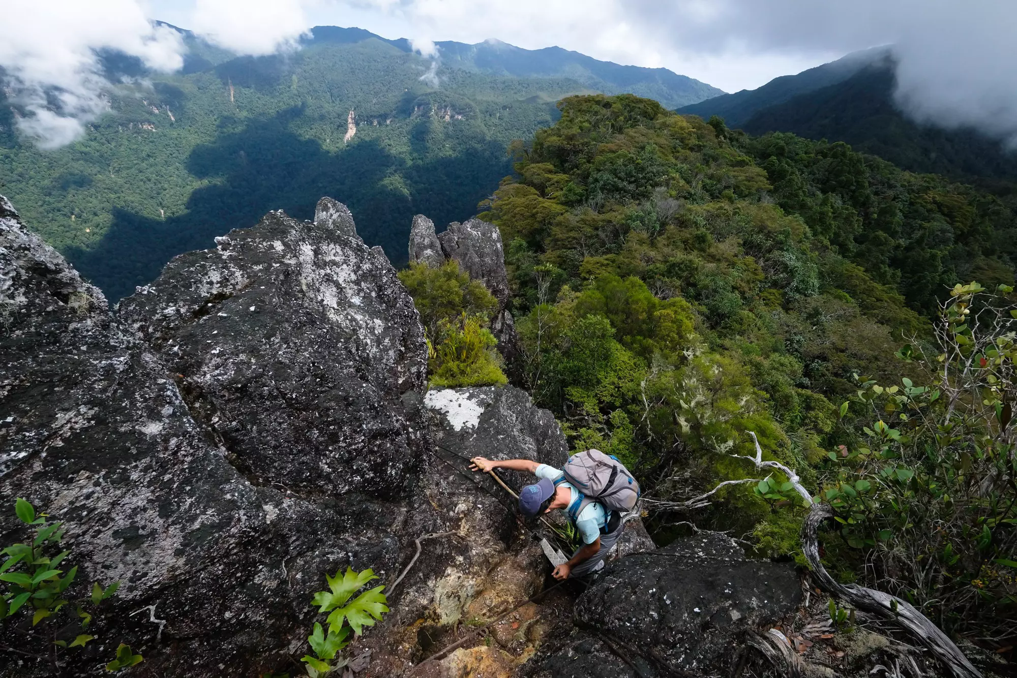 A hiker seen from above scaling a rock face on a mountain covered with tropical vegetation, with wispy clouds in the distance