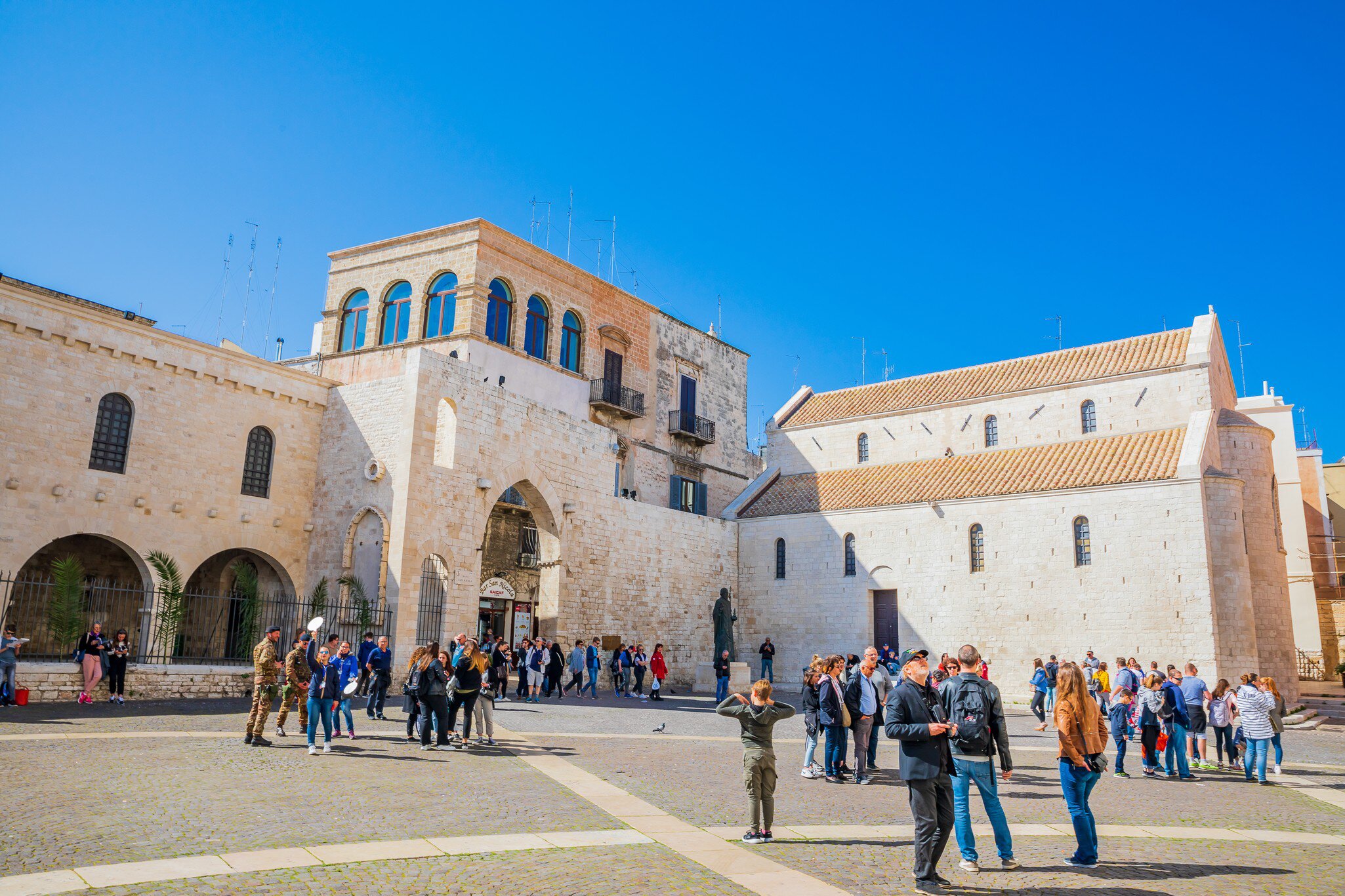 Pale limestone church with tiled roofs beside an arched gatehouse, tourists crossing a sunny paved square