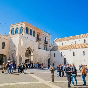 Pale limestone church with tiled roofs beside an arched gatehouse, tourists crossing a sunny paved square