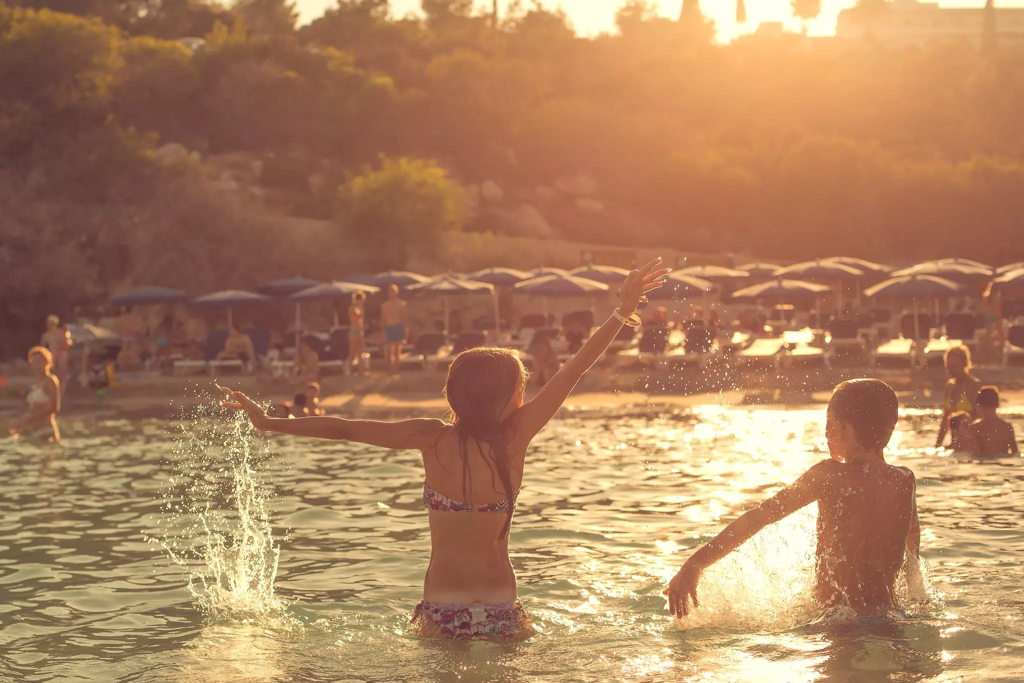 Two pre-teen children splash together in the sea as the sun sets over a beach lined with sunloungers