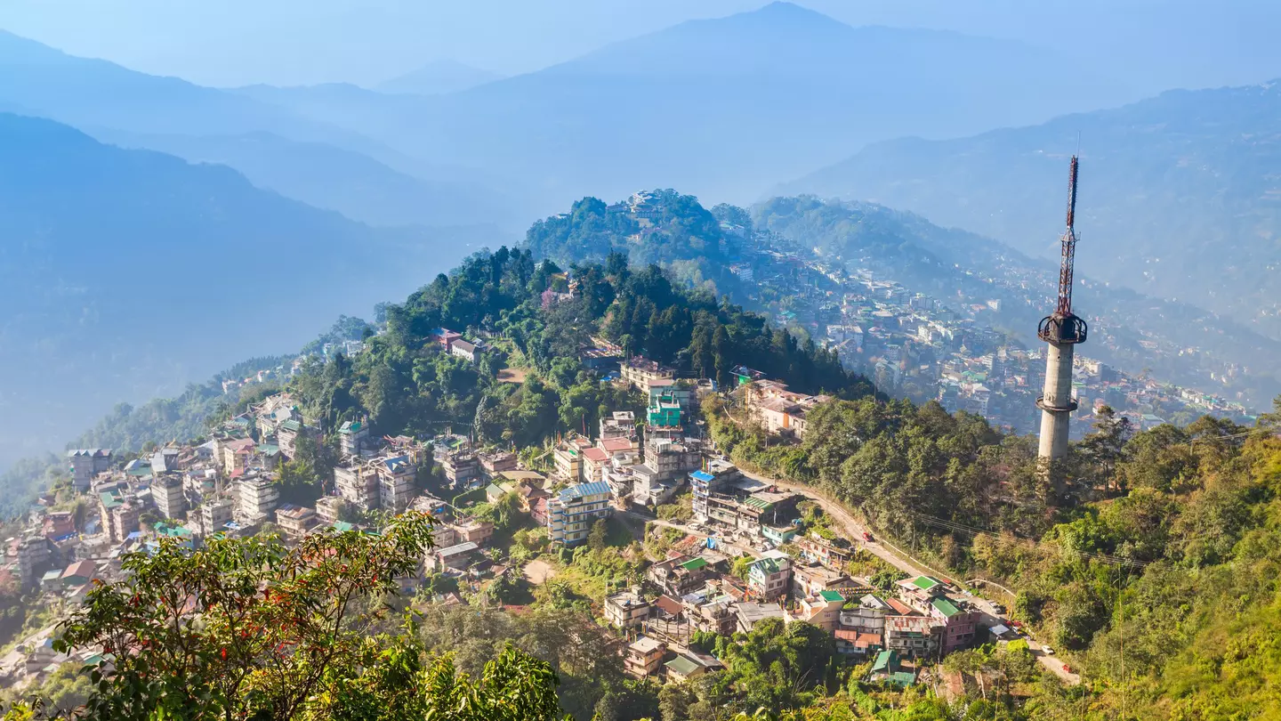 A small city spread out on hillsides and mountain peaks surrounded by green foliage on a sunny day.