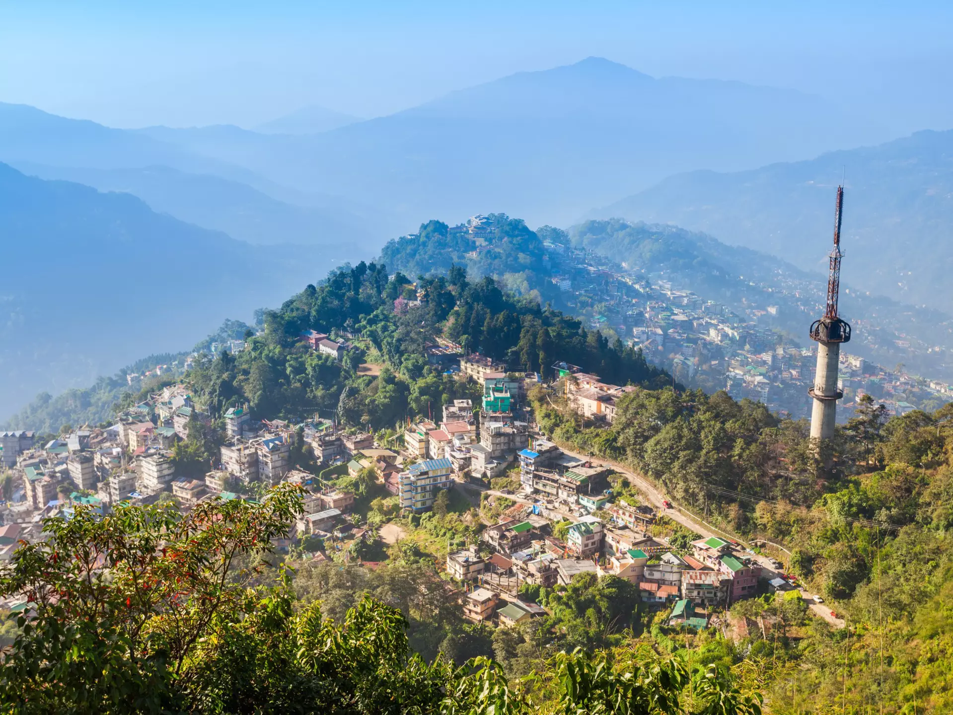 A small city spread out on hillsides and mountain peaks surrounded by green foliage on a sunny day.