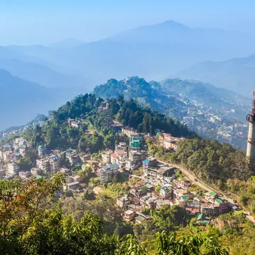 A small city spread out on hillsides and mountain peaks surrounded by green foliage on a sunny day.