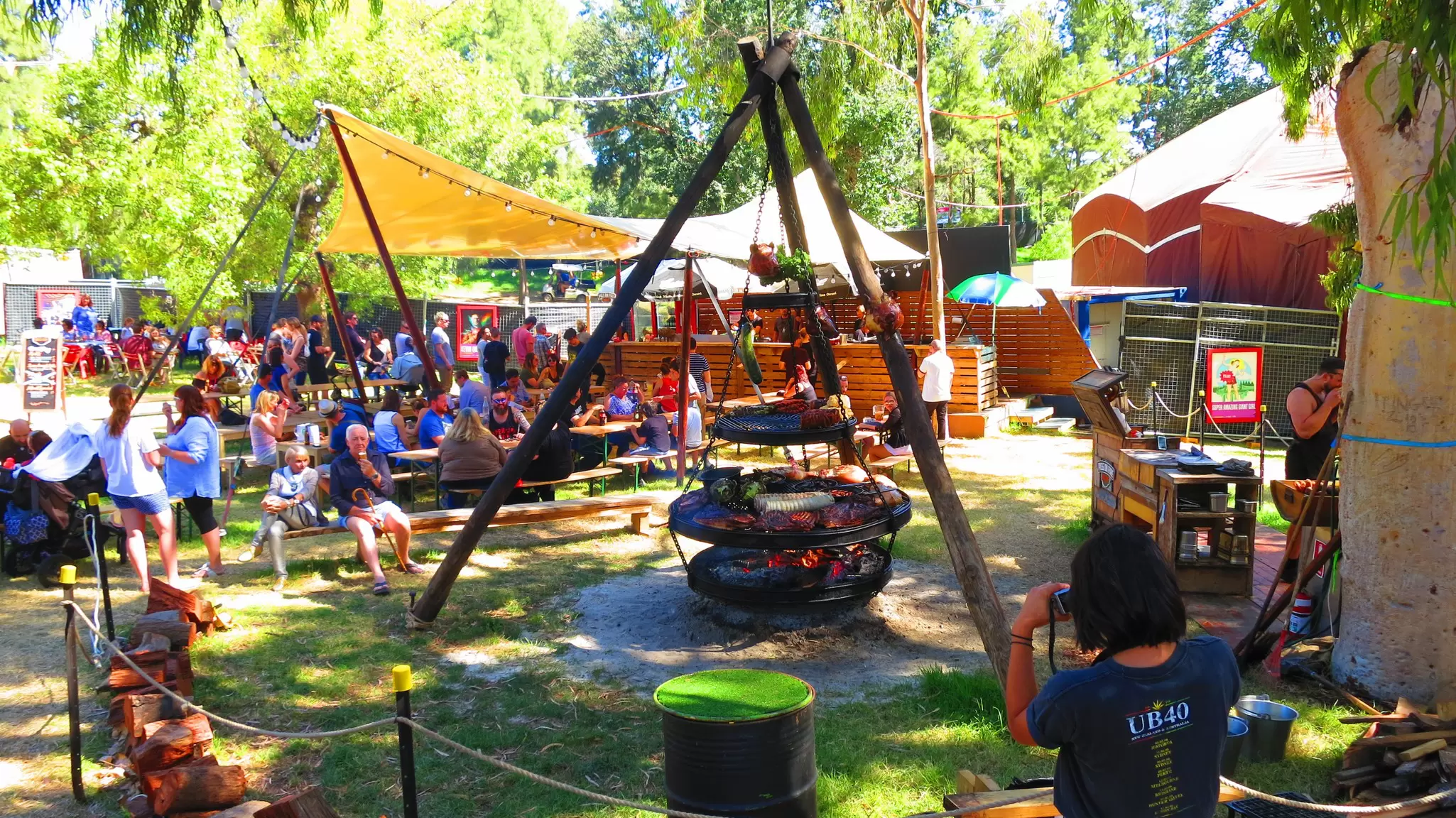 Colourful people sitting around a wood fired BBQ with stalls behind them at the Adelaide Fringe Festival in South Australia. 