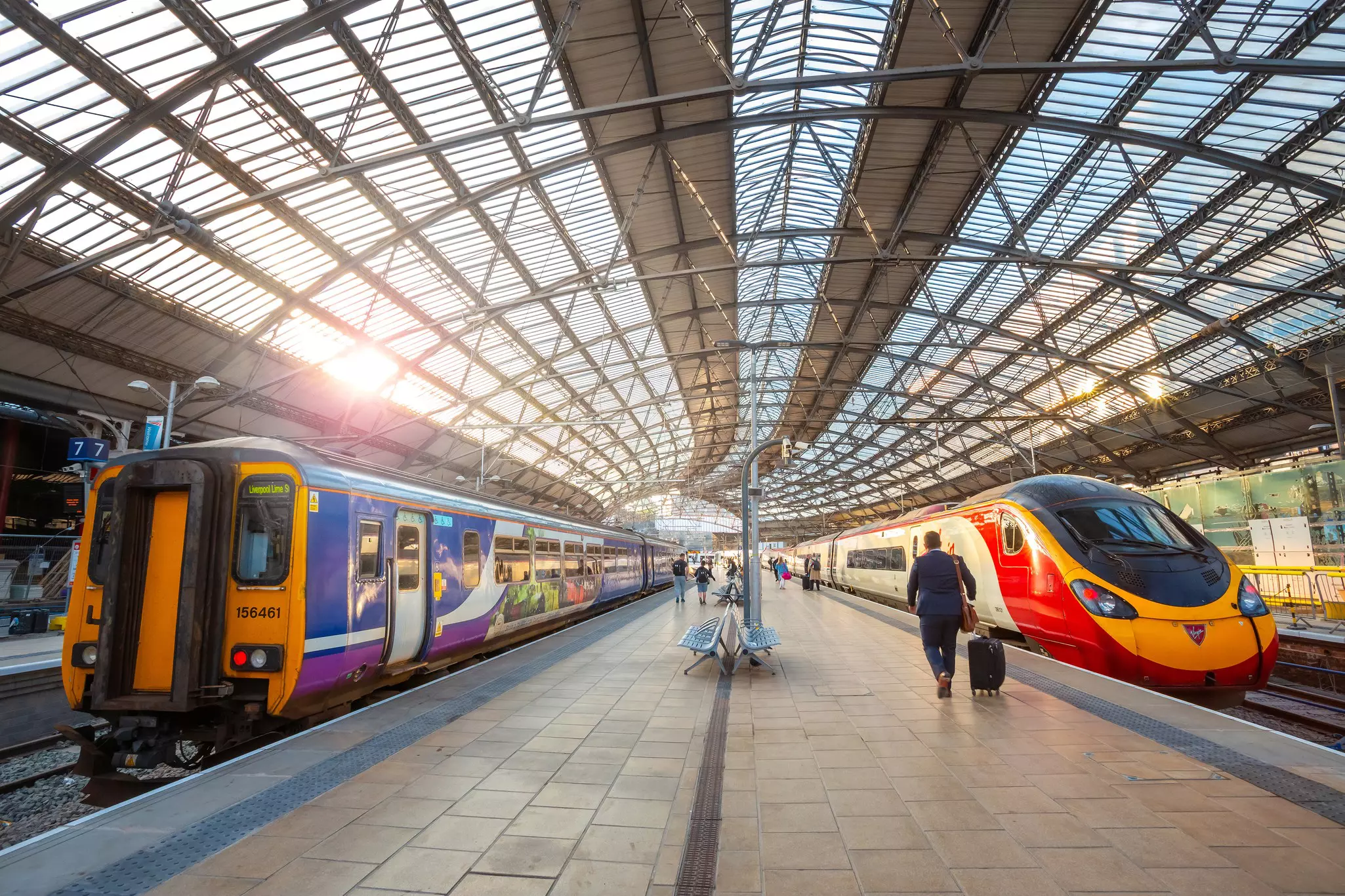 Two trains stand either side of a central platform in a station with a curved glass and iron roof.