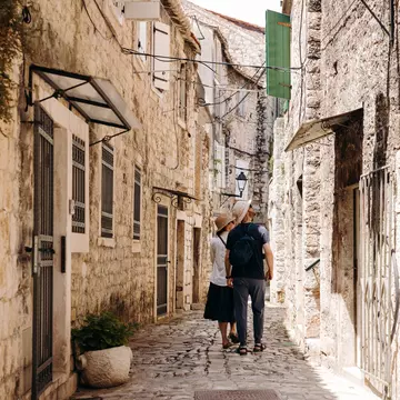 People walk on a narrow street looking up at the stone buildings.