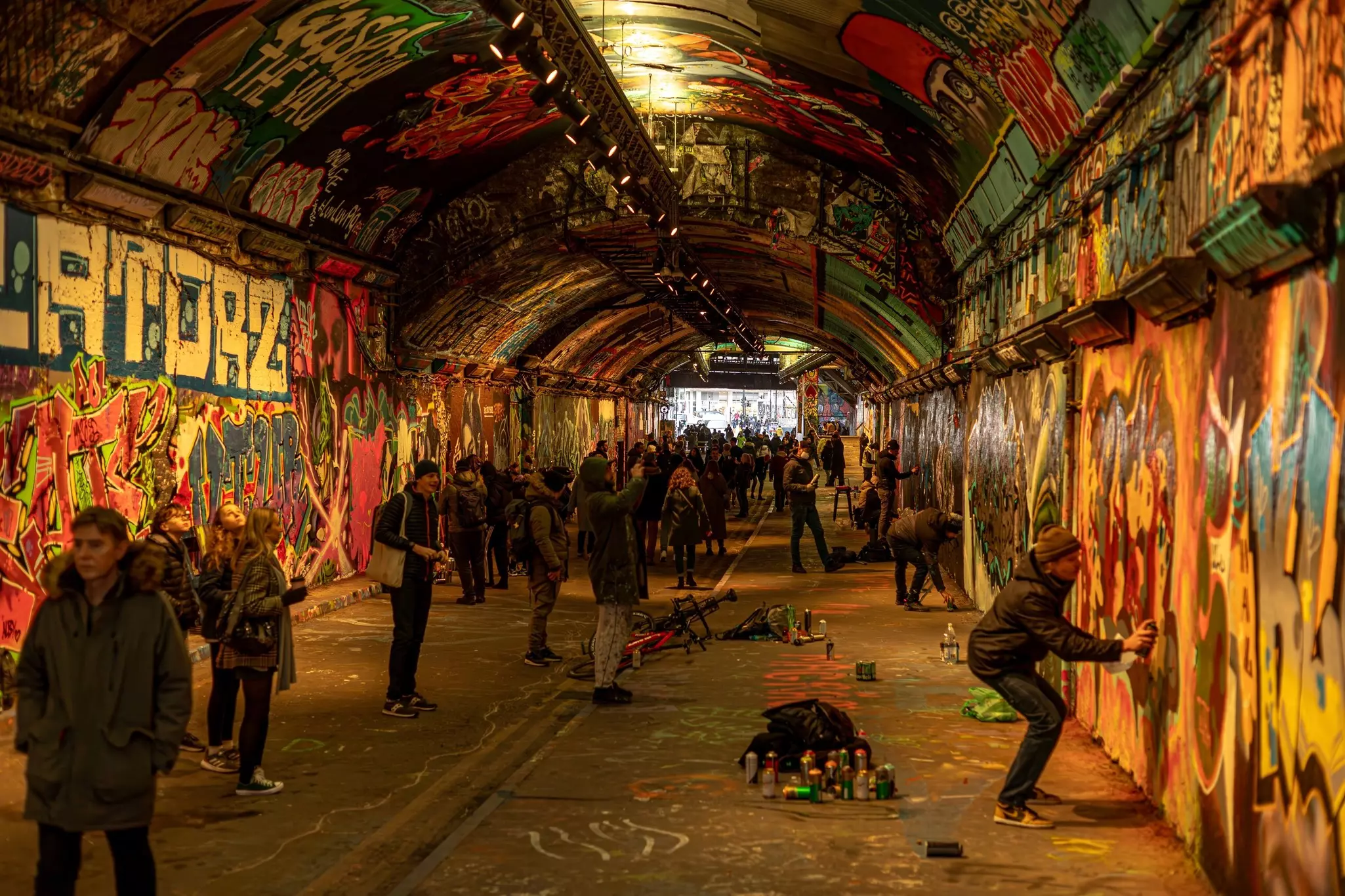 People walking and spraypainting a long tunnel filled with graffiti.