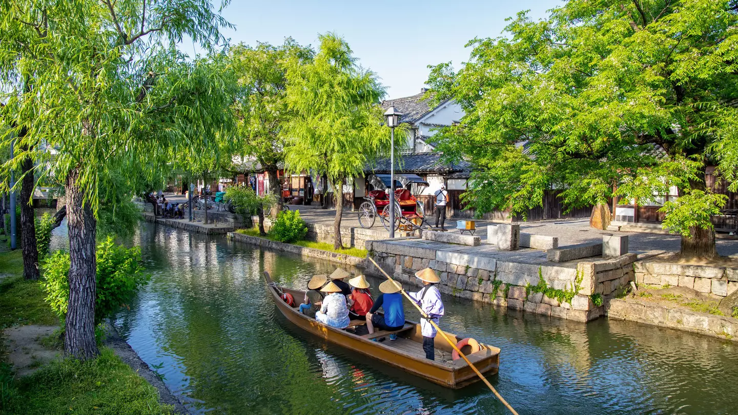 People wearing conical hats ride in a boat on a narrow waterway through a town in Japan.