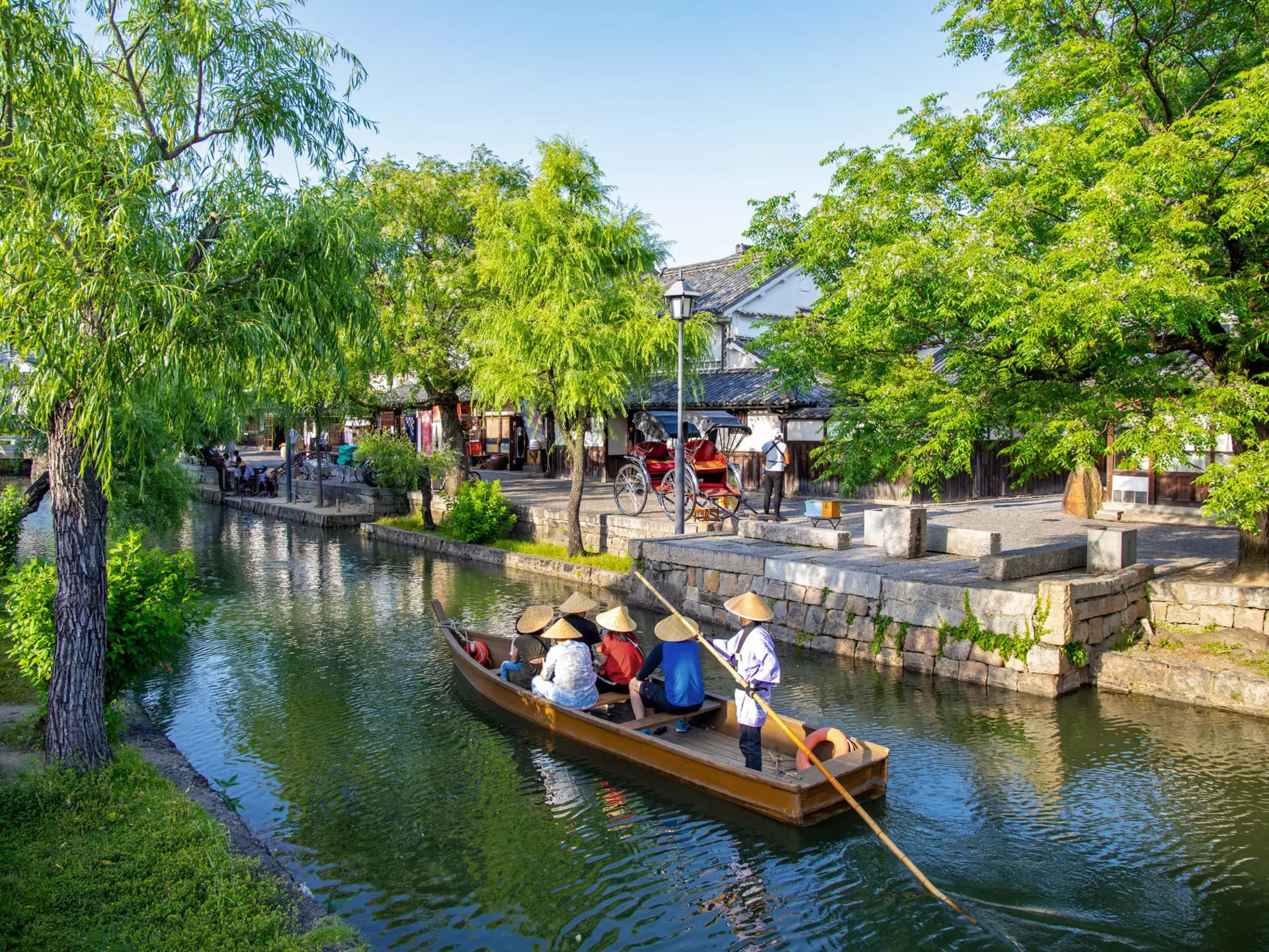 People wearing conical hats ride in a boat on a narrow waterway through a town in Japan.