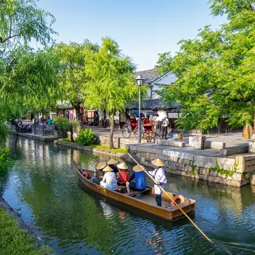 People wearing conical hats ride in a boat on a narrow waterway through a town in Japan.