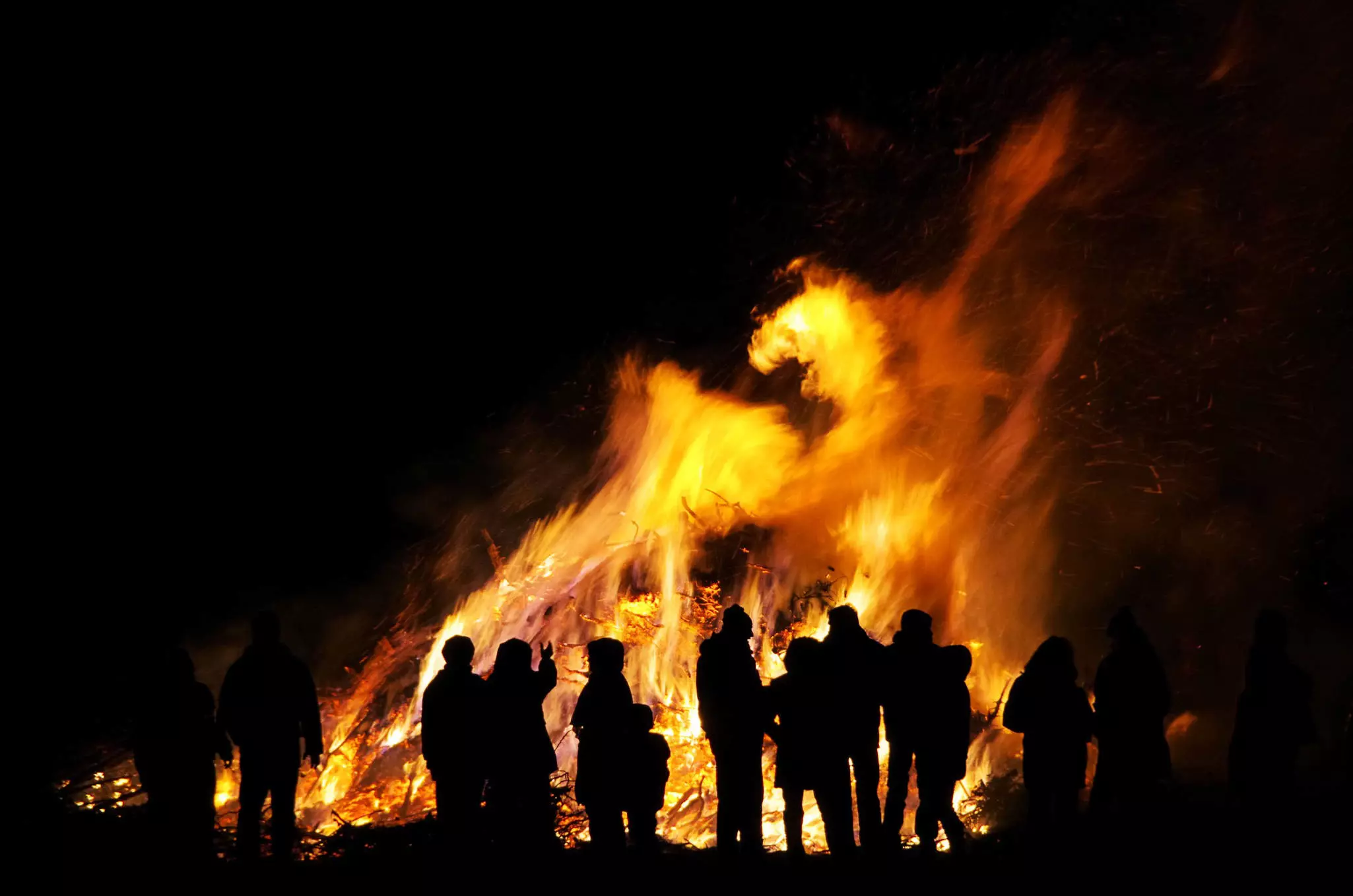 Silhouettes of people standing around a large bonfire that towers above them.