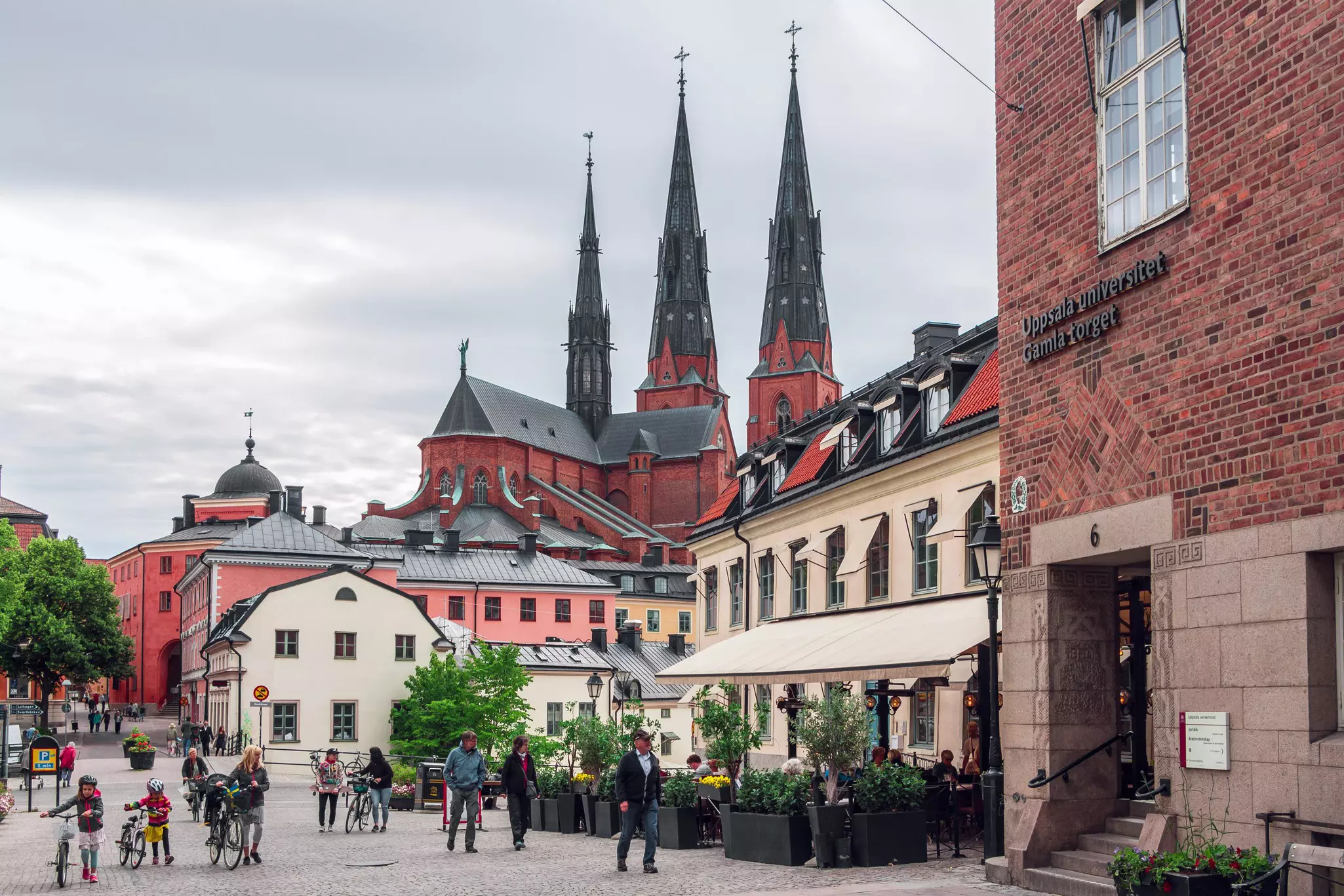 People walk and ride bikes on a pedestrianized street in a city. A tall church with three spires is seen in the background.