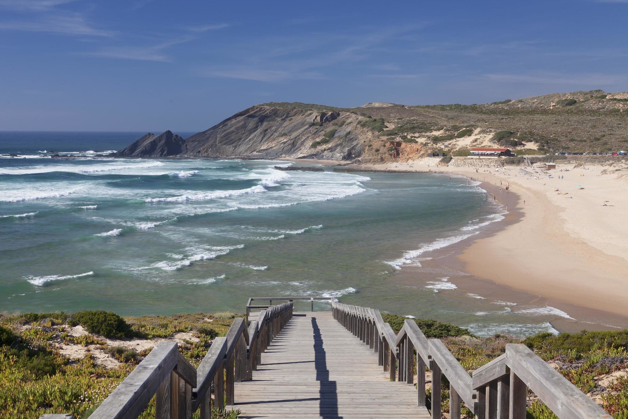 A wooden staircase down to a long, sandy beach and the ocean, with cliffs in the background.