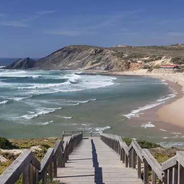 A wooden staircase down to a long, sandy beach and the ocean, with cliffs in the background.