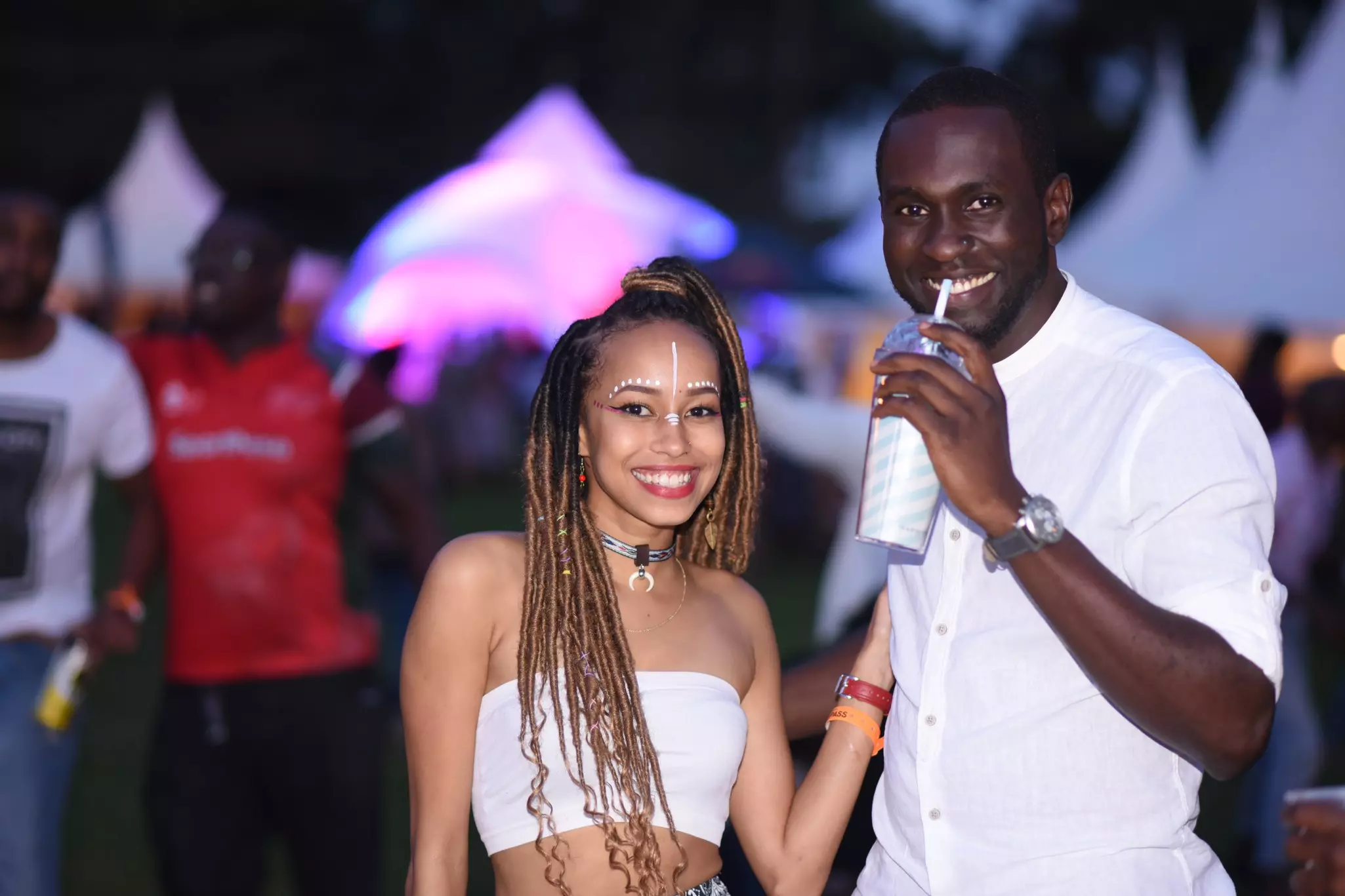 Two young people smile as one sips a drink at an evening outdoor concert.