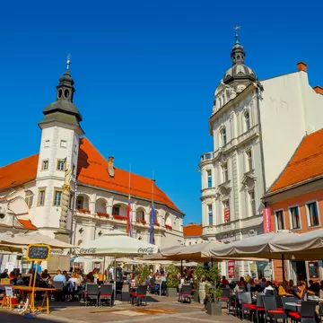 A sunny town square in Maribor with a white town hall and bell tower, orange roofs and people at café tables under umbrellas.