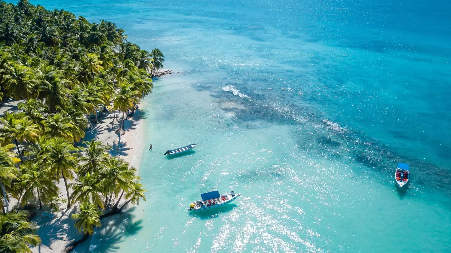 Boats in clear turquoise water offshore of a sandy beach with palm trees.