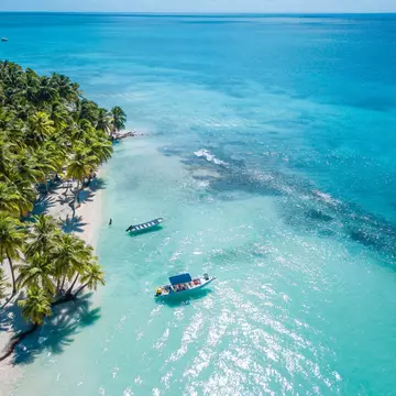 Boats in clear turquoise water offshore of a sandy beach with palm trees.