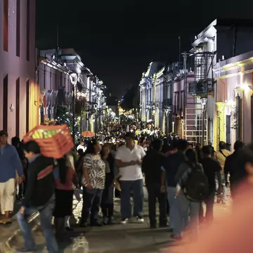 We see a crowded night street in downtown Oaxaca City in Mexico, during the celebration time and annual Guelaguetza celebration. the street is very crowded; the focus is in the background; the front people are in soft focus.