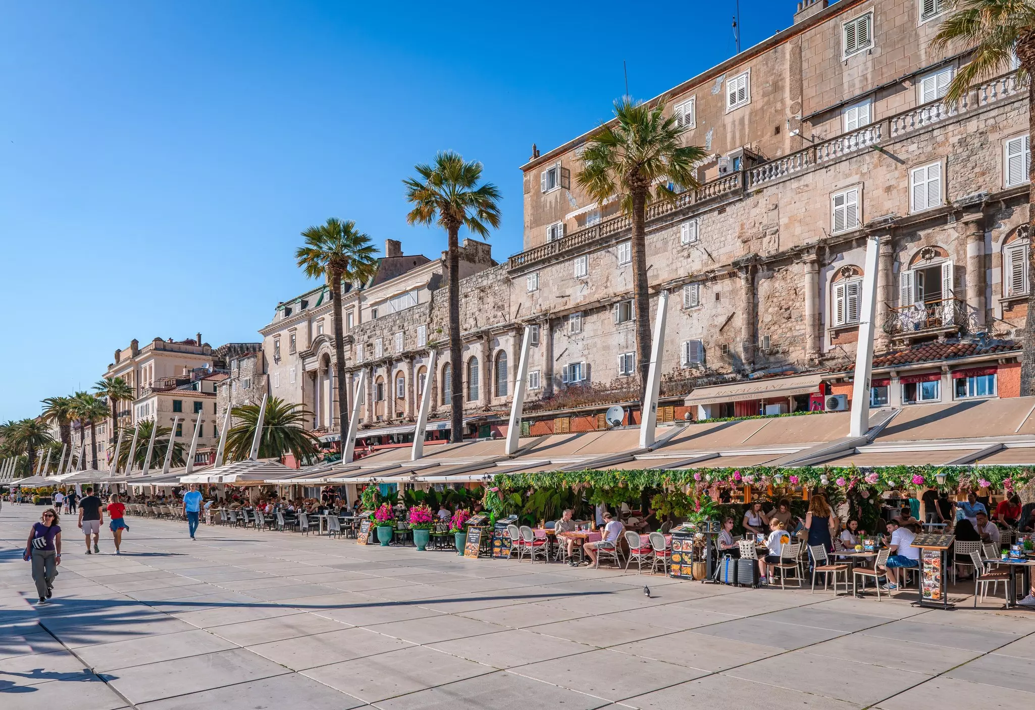 People gathered at cafe tables under awnings that line a seafront walkway.