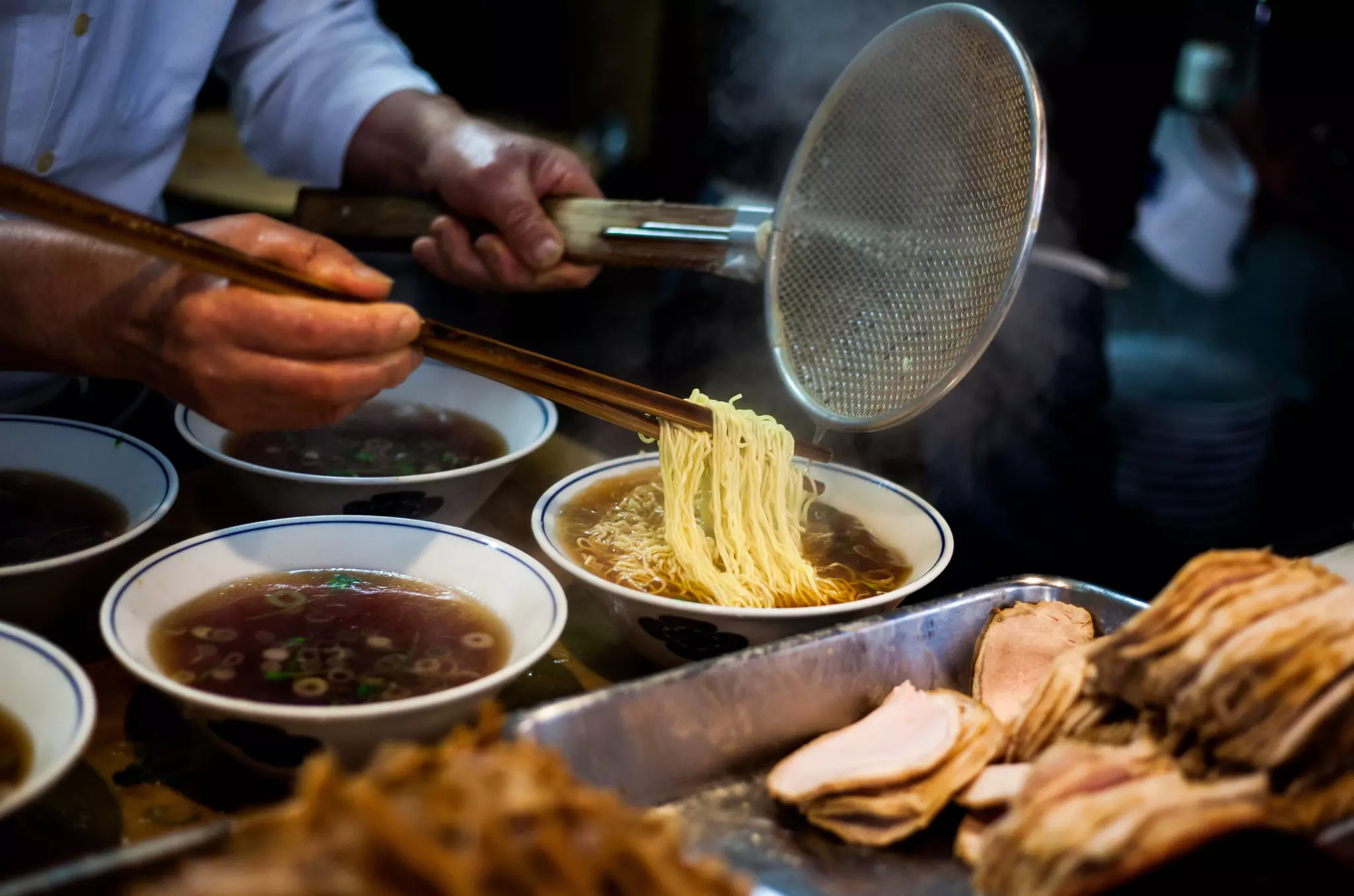 Hands preparing steaming ramen (soba) while holding long chopsticks and metal strainer.