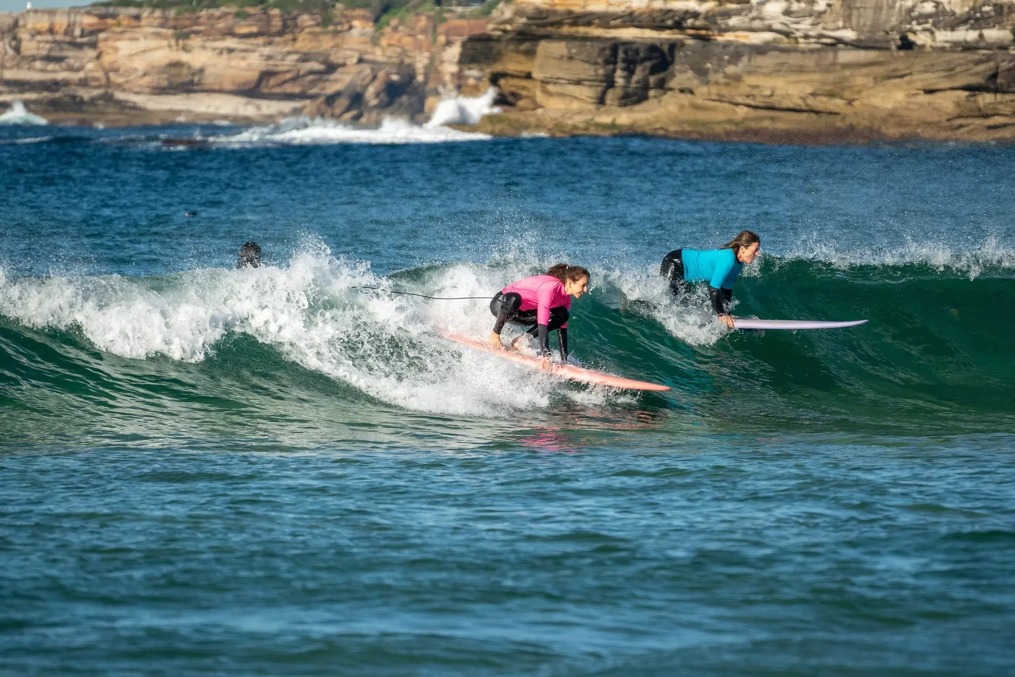Two women about to catch a wave while surfing on the ocean with a rock wall in the distance on a sunny day.