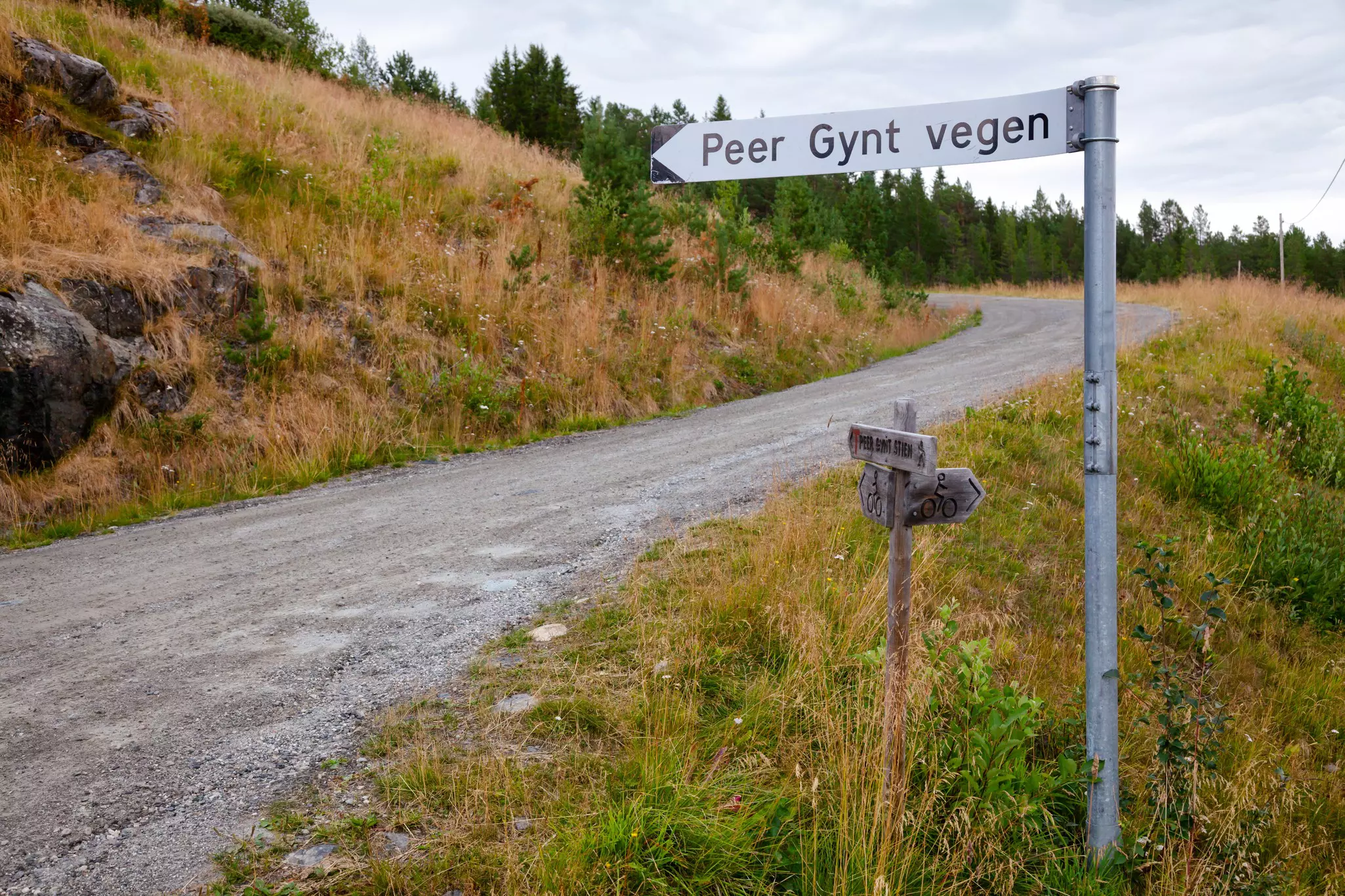 An arrow-shaped sign points toward a narrow dirt road on a hillside in the Norwegian countryside.