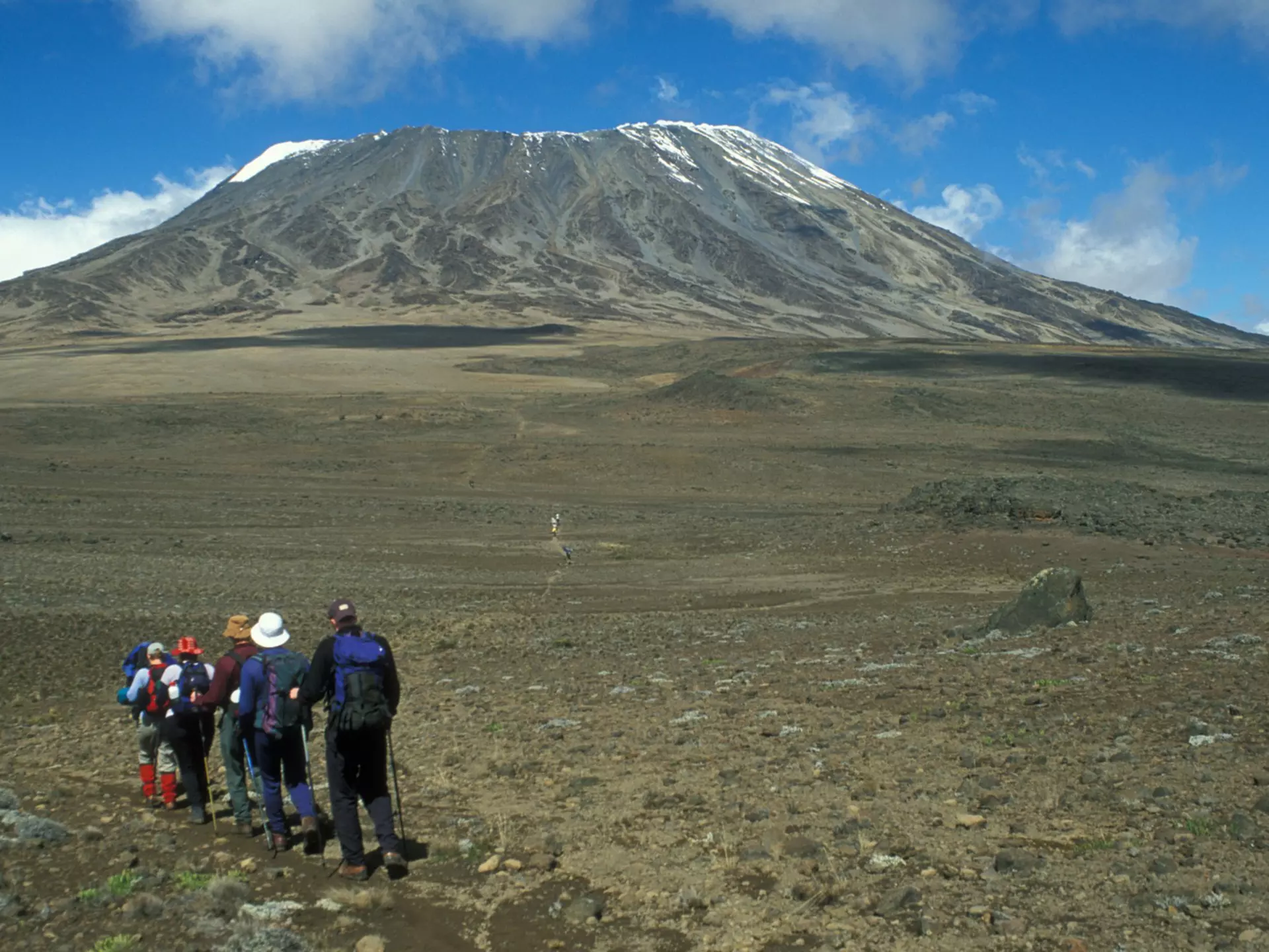 A group of hikers walk in a line across a barren field, with the peak of a mountain rising in the near distance.