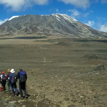 A group of hikers walk in a line across a barren field, with the peak of a mountain rising in the near distance.