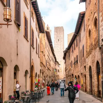 People stroll a narrow cobbled street in San Gimignano, Italy, between tall brick and stone buildings, a square tower ahead.