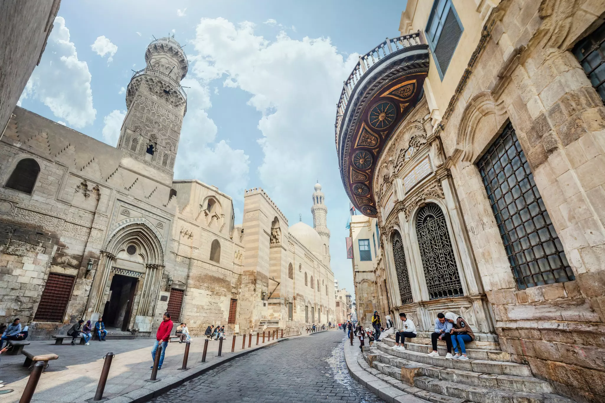 People sitting on steps and benches along a narrow street lined with ancient buildings and a minaret rising above.