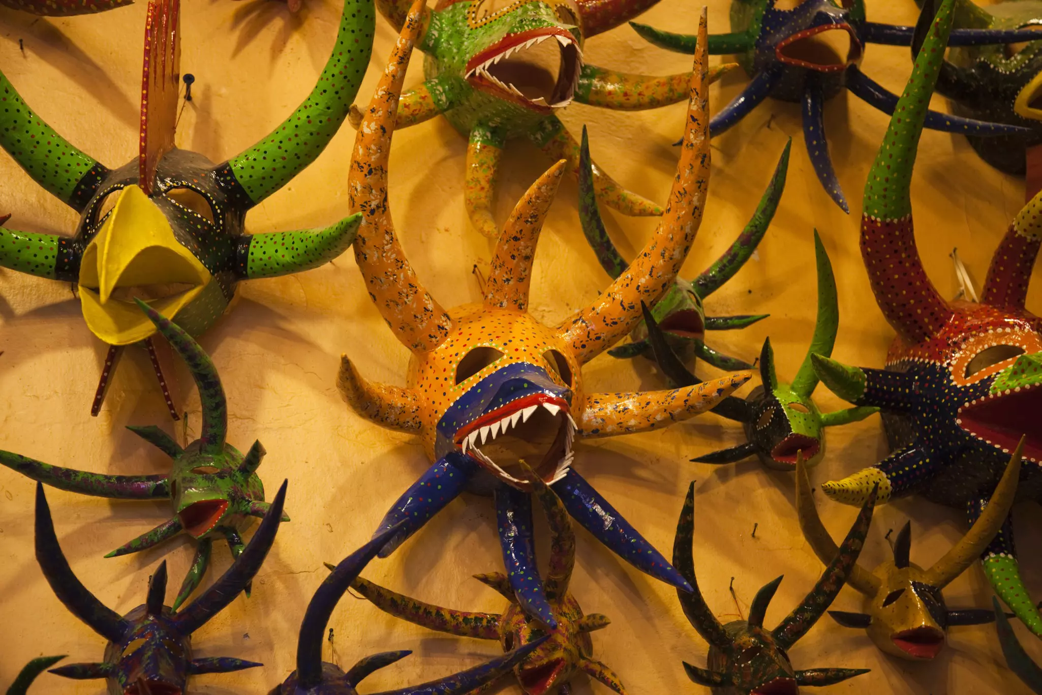 A collection of colorful masks with large horns, some with sharp teeth and another with a large beak are hung on a yellow wall.