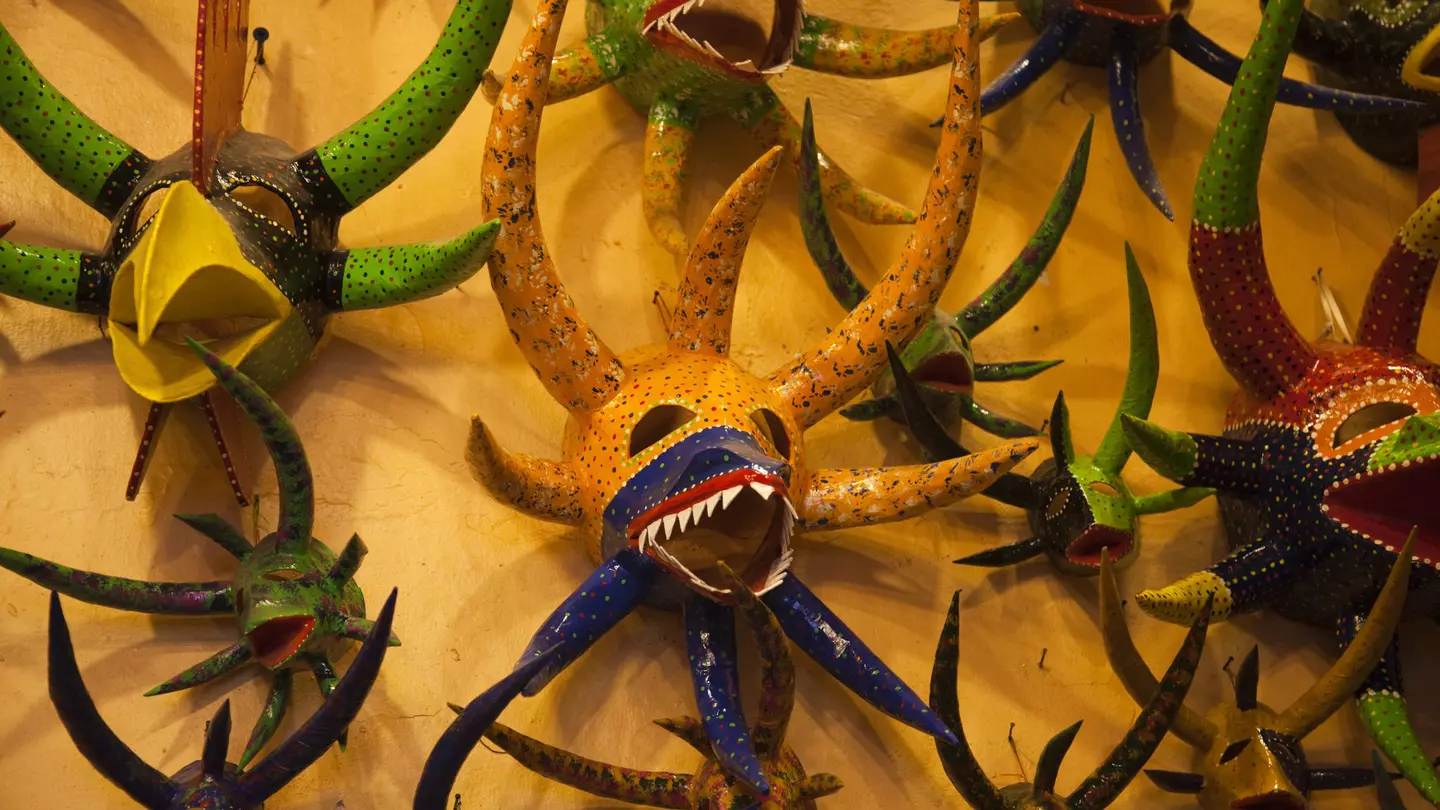 A collection of colorful masks with large horns, some with sharp teeth and another with a large beak are hung on a yellow wall.