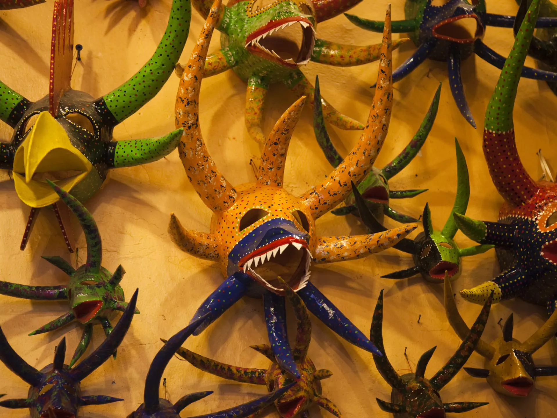 A collection of colorful masks with large horns, some with sharp teeth and another with a large beak are hung on a yellow wall.