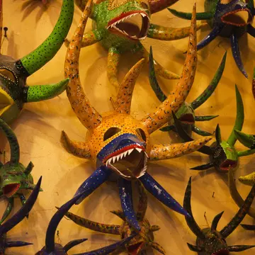 A collection of colorful masks with large horns, some with sharp teeth and another with a large beak are hung on a yellow wall.