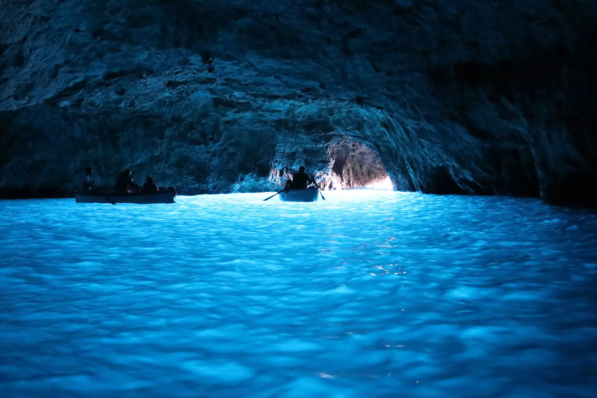 Two small boats atop bright blue water in a cave with sunlight peeking in from an arched entry.