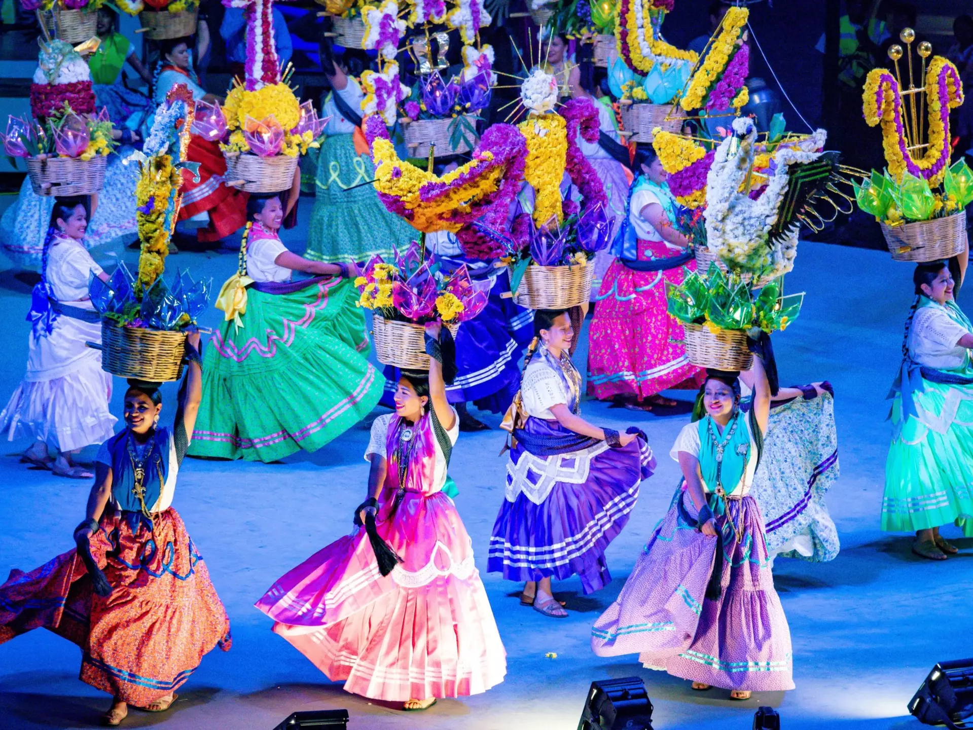 A group of young females in colorful dresses dancing with baskets full of flowers on a stage.