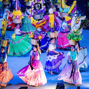 A group of young females in colorful dresses dancing with baskets full of flowers on a stage.
