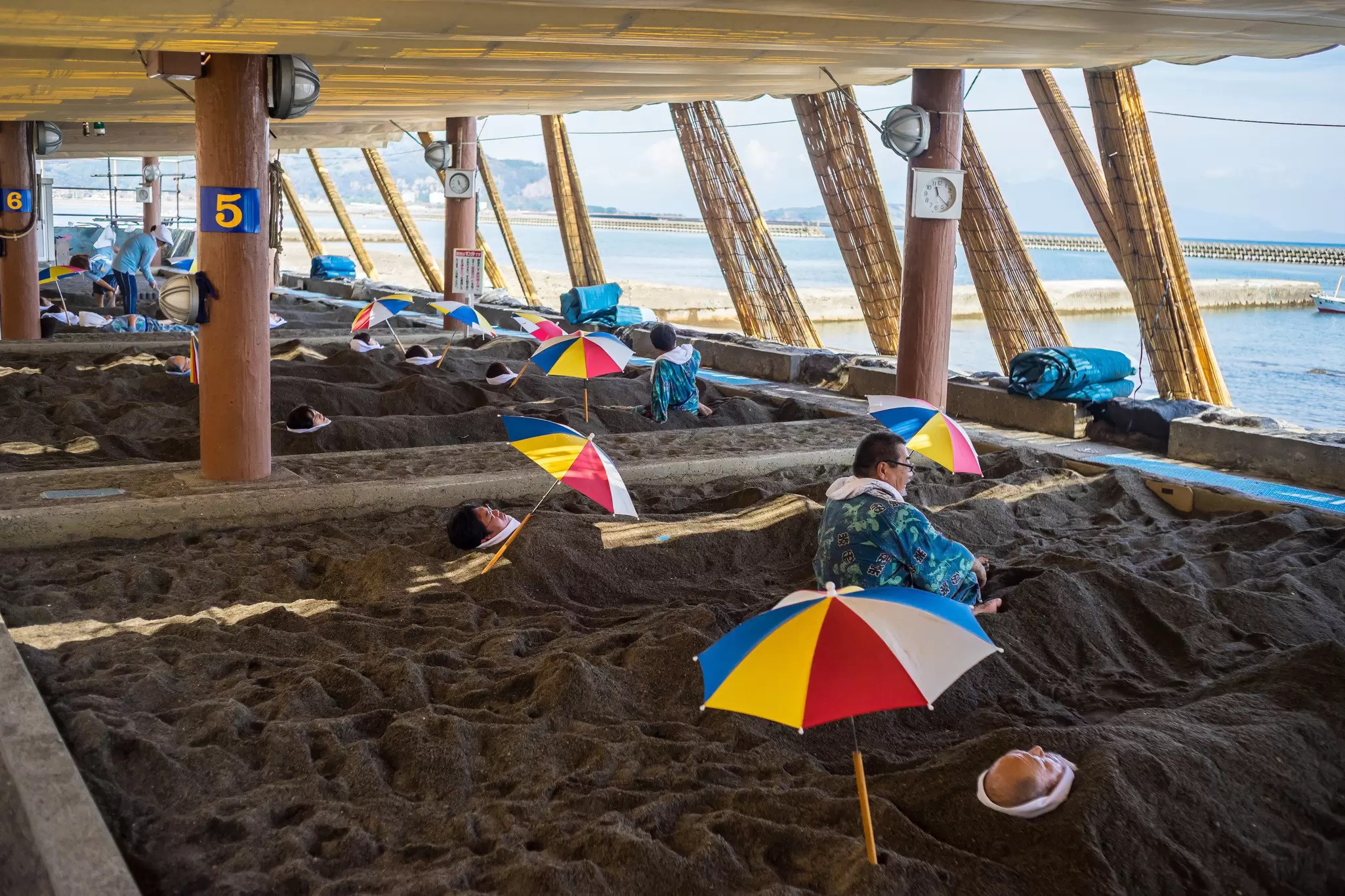 People covered in sand under a wooden structure by the water.