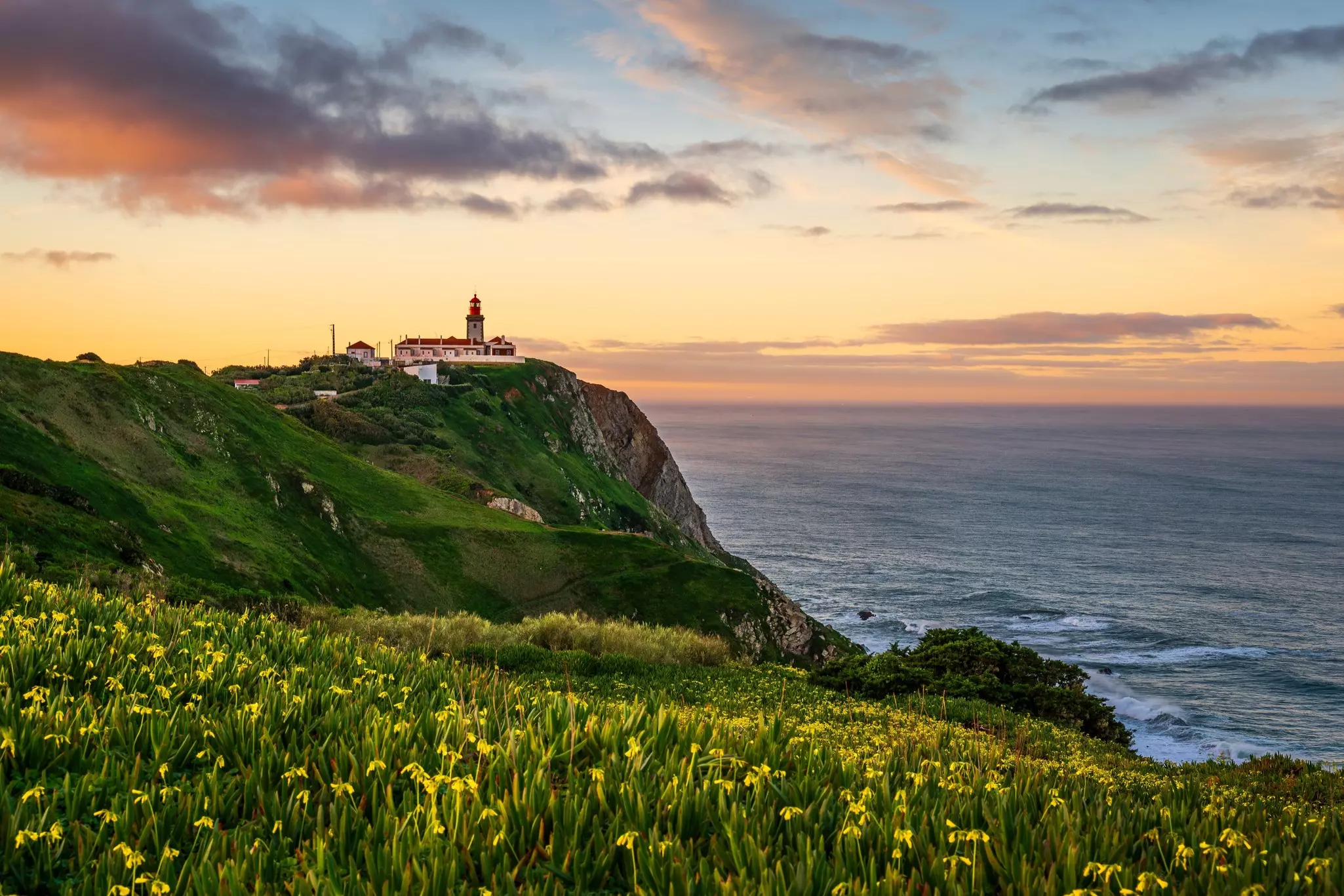 A lighthouse as the edge of a cliff by the sea at sunset.