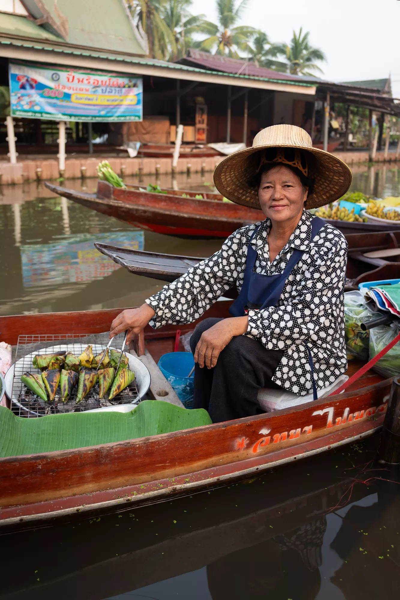 A woman in a straw hat sits on a boat in a floating market selling snacks being heated on a grill.