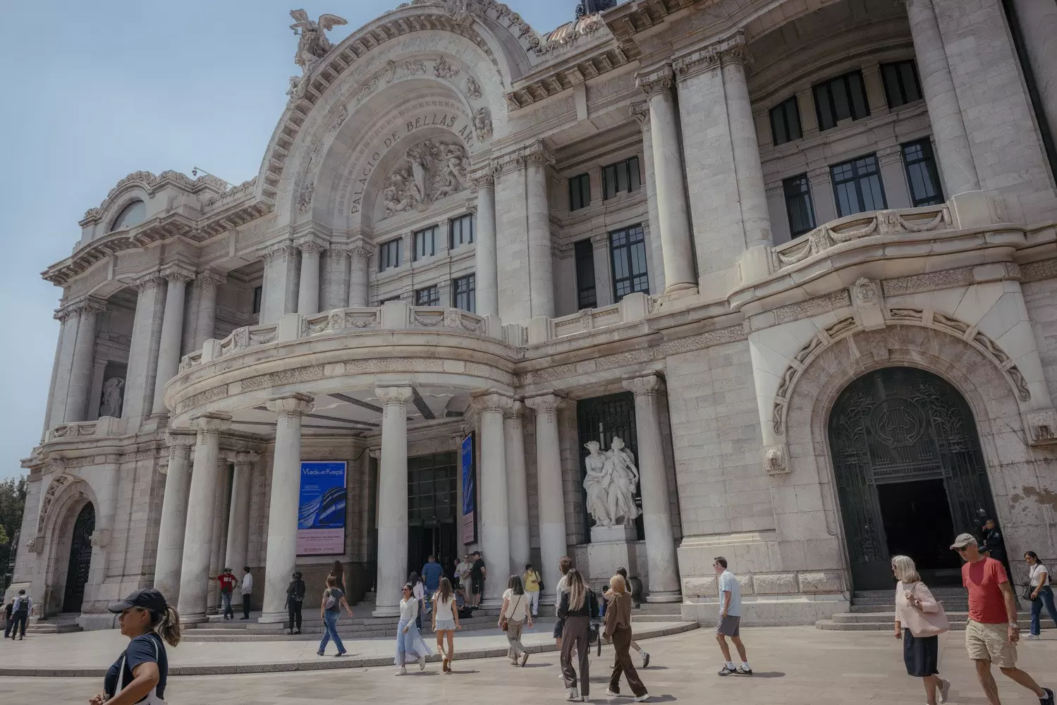 People walk toward a large white building in Mexico City with archways, a columned portico entryway and intricate details on the roofline.