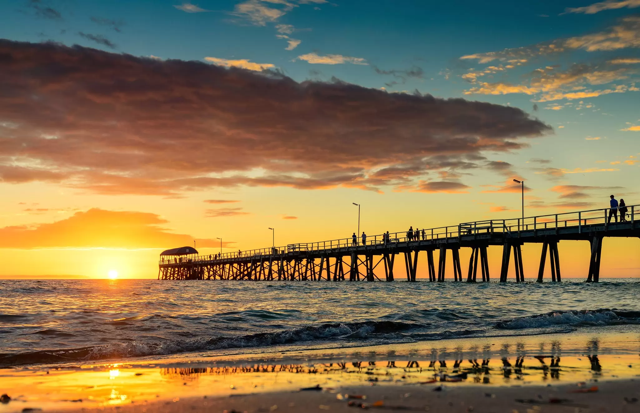 People walking on the Henley Beach Jetty at sunset, shot taken from beach with water reflecting the sunset sky