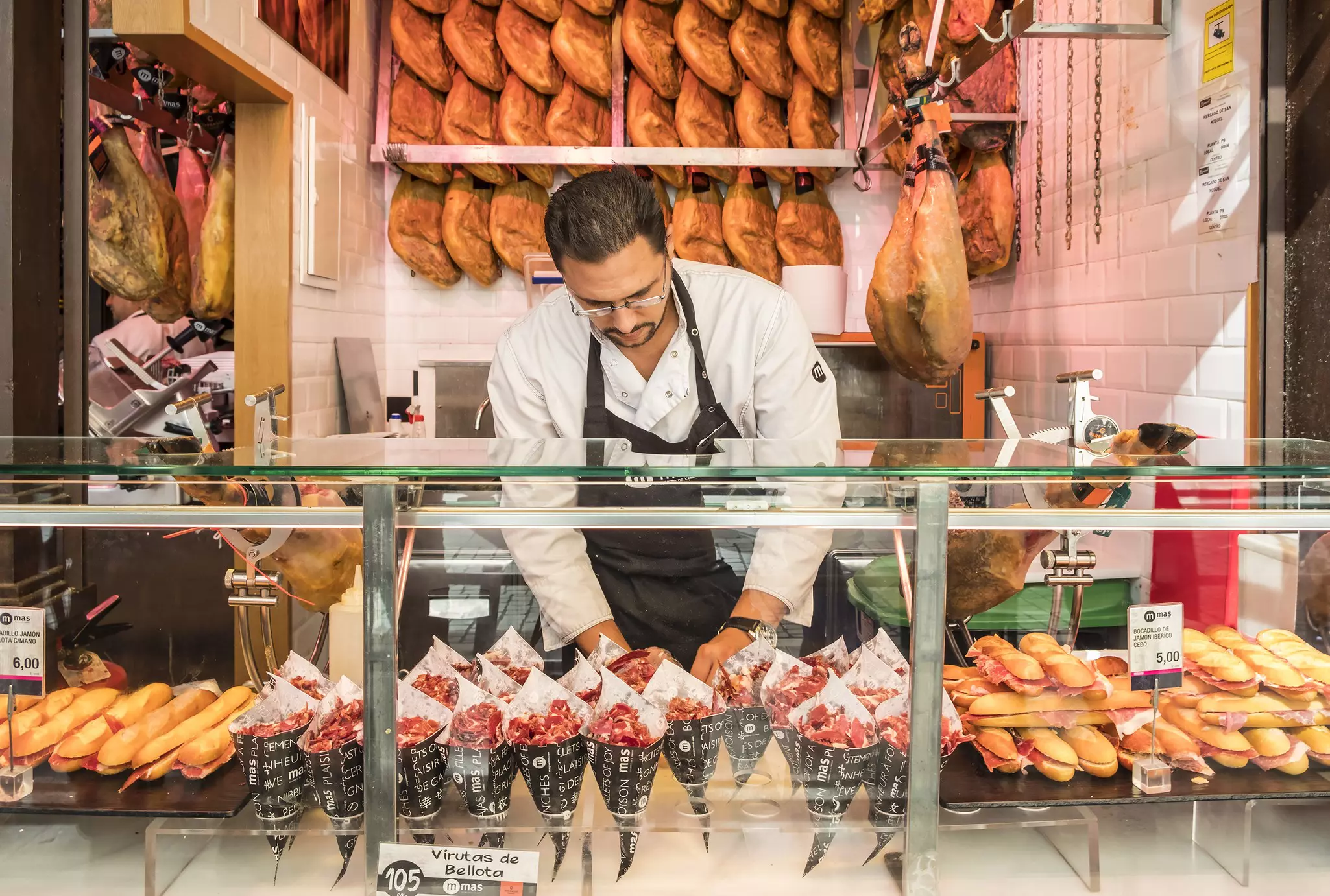 A man at a food stall in a market. On display are cones filled with ham, bread rolls filled with ham and cheese and behind him, hanging on the wall, are legs of ham.