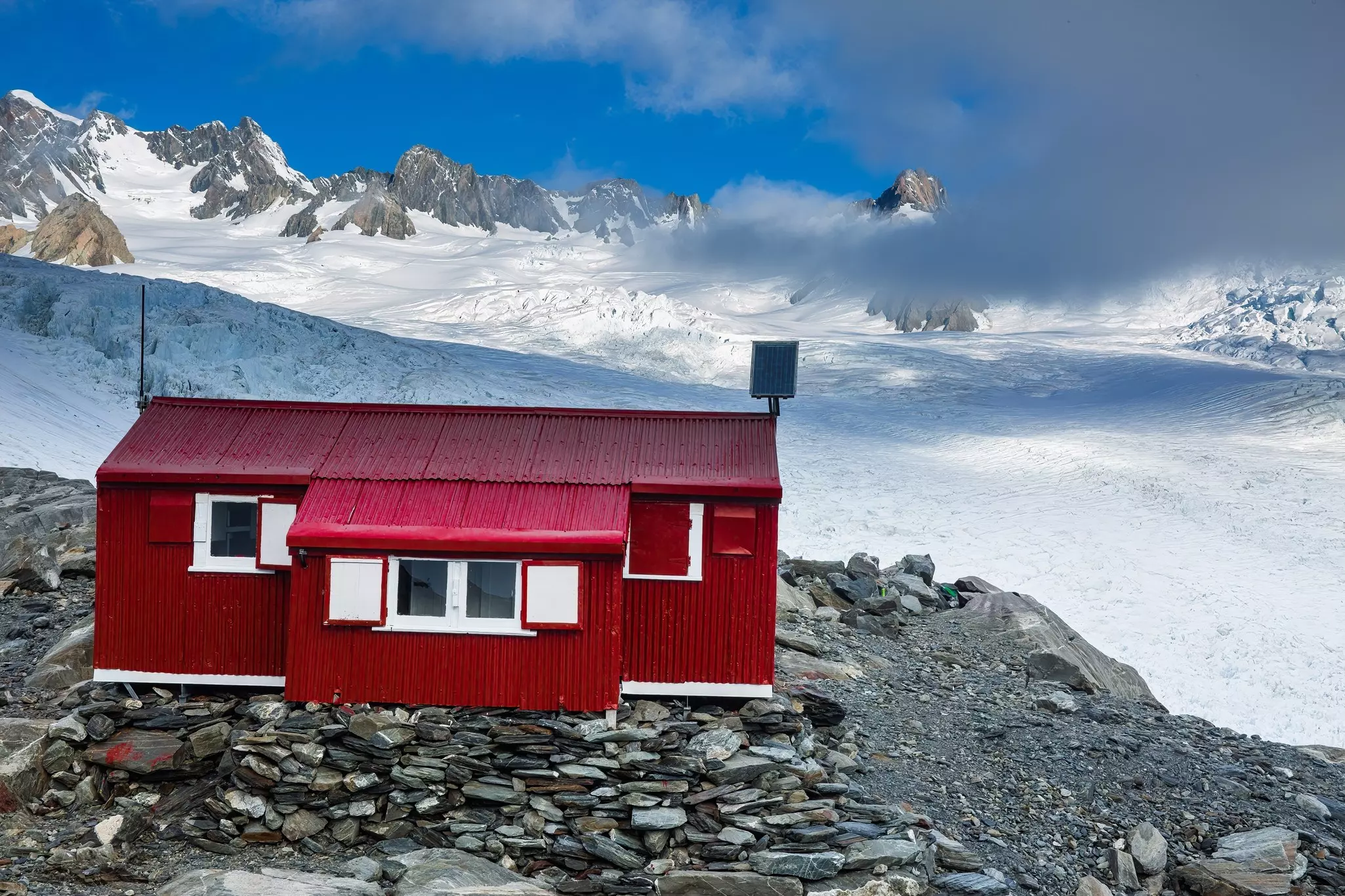 A small red hut with corrugated cladding on a rocky precipice in a snowy mountainous area.