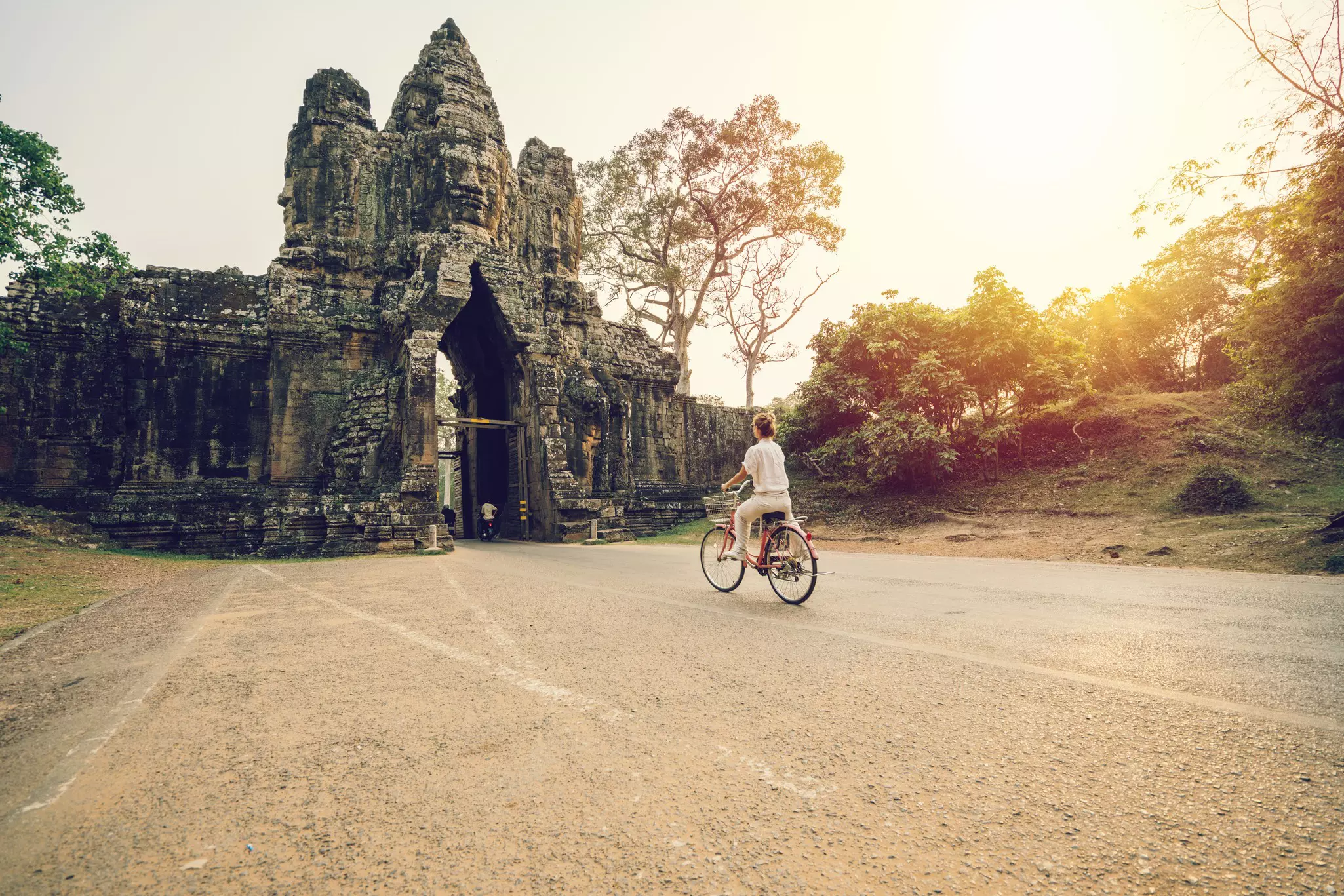 Three tourists, each wearing helmets, stand straddling mountain bikes; their backs are to the camera and they are facing some ancient ruins and crowds of people. The man in the middle is using his phone to take a picture.