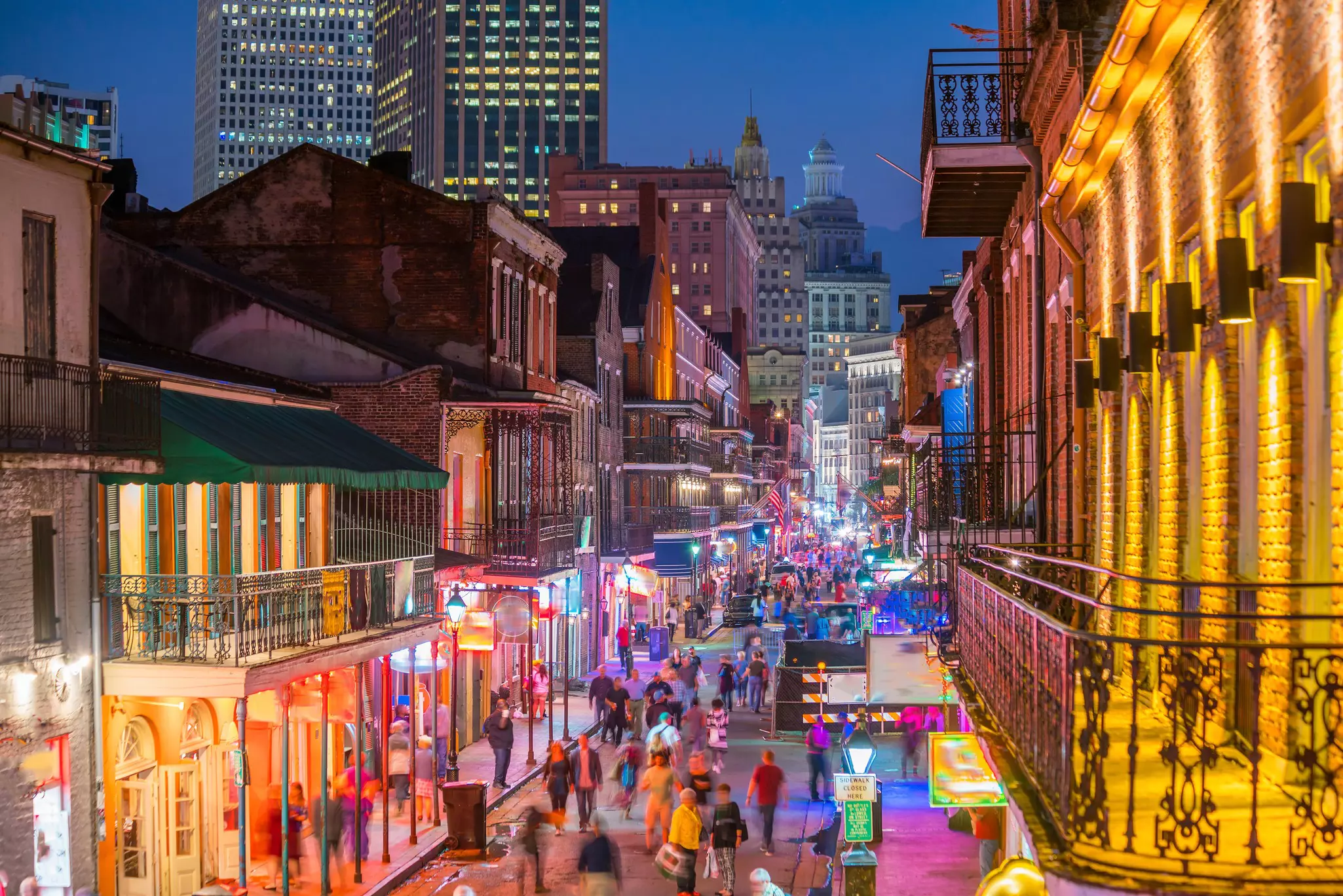 A pedestrianized street is lit by colored lights and has numerous bars. Skyscrapers are in the distance behind the historic buildings of the neighborhood.