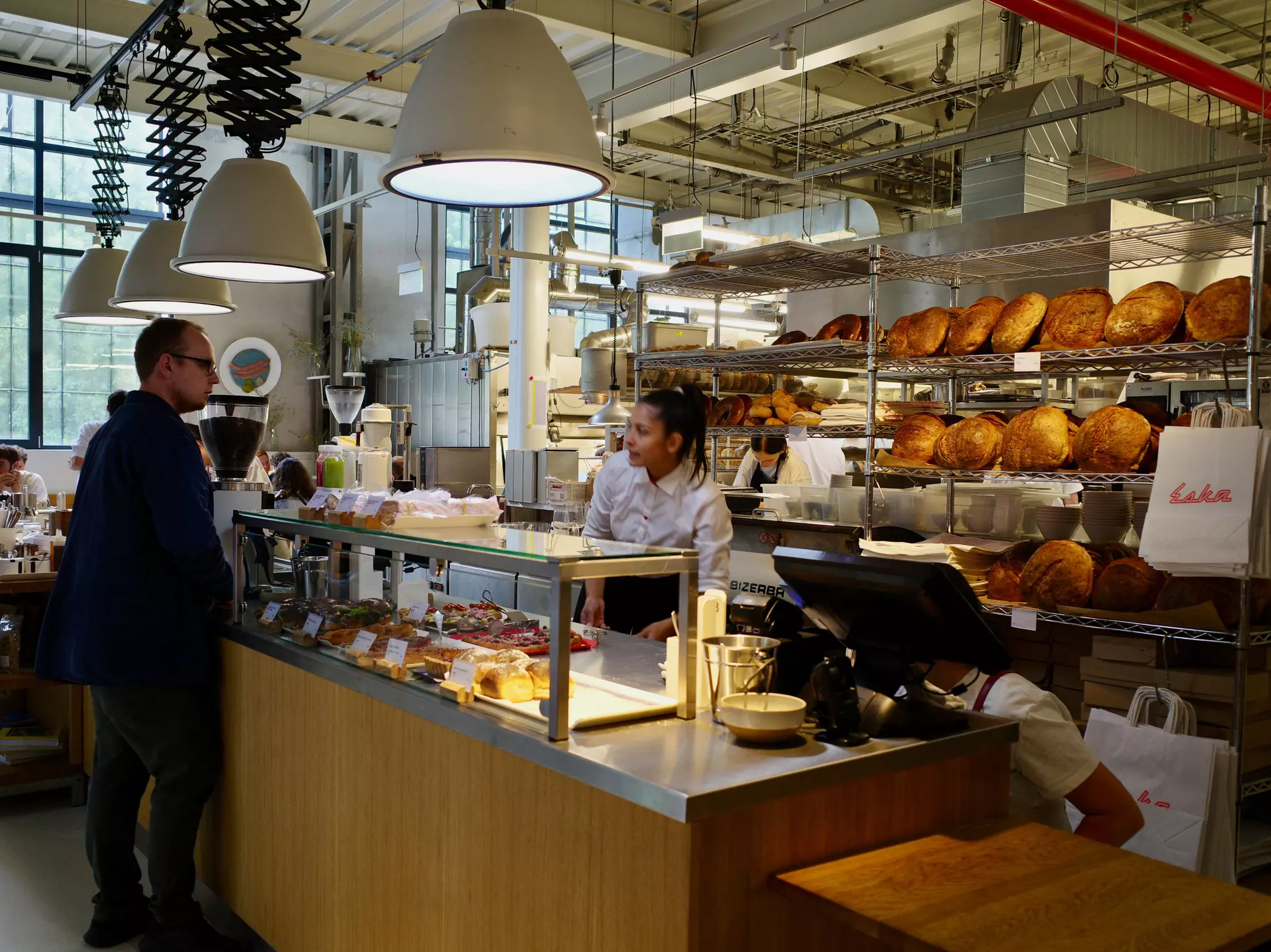 A person making an order at a bakery counter. Rows of large, full breads line shelves behind the counter.