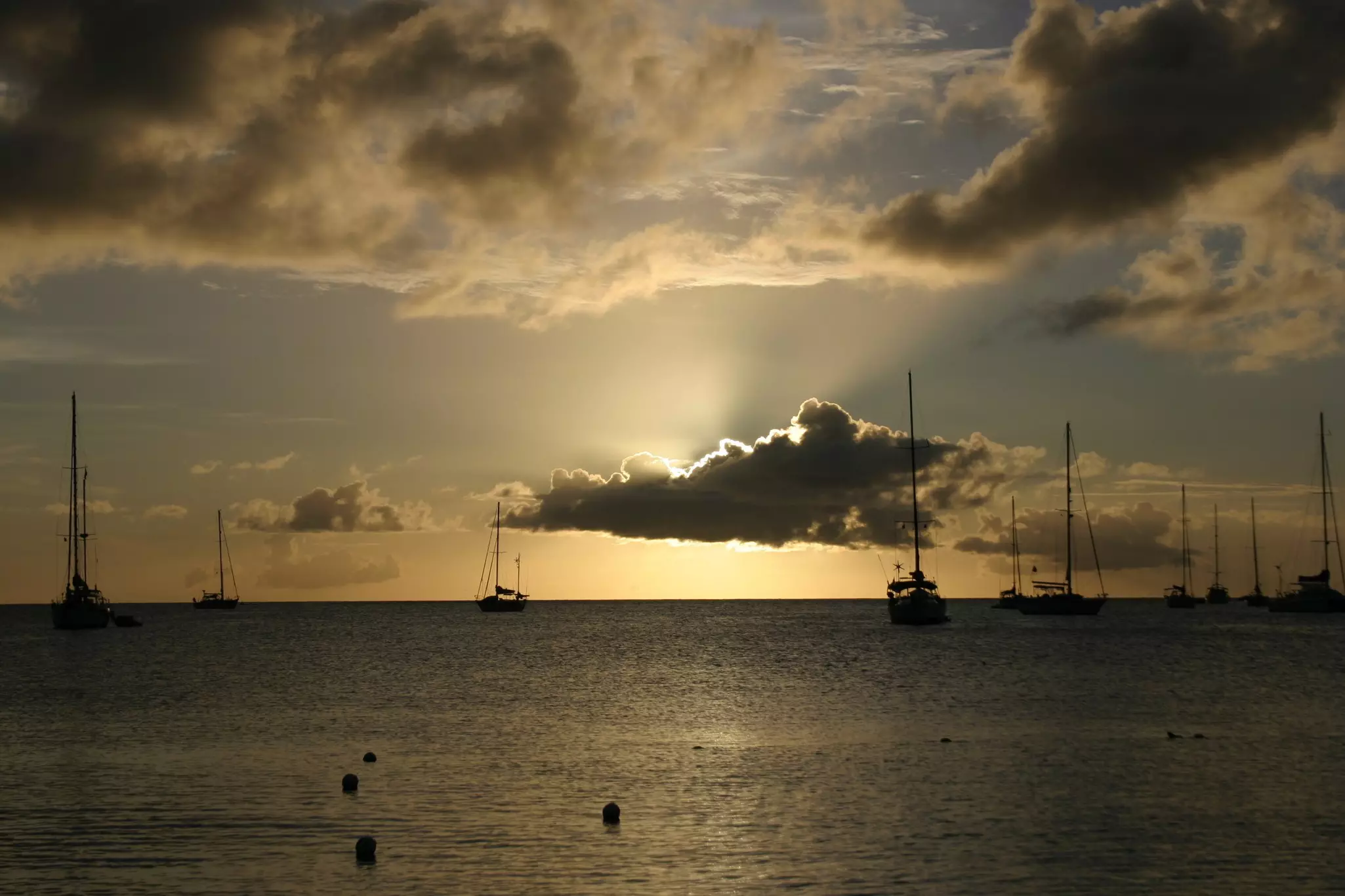 A cloud blocks the setting sun over the water, making for dramatic shadow effects. Multiple sailboats are moored in the water.
