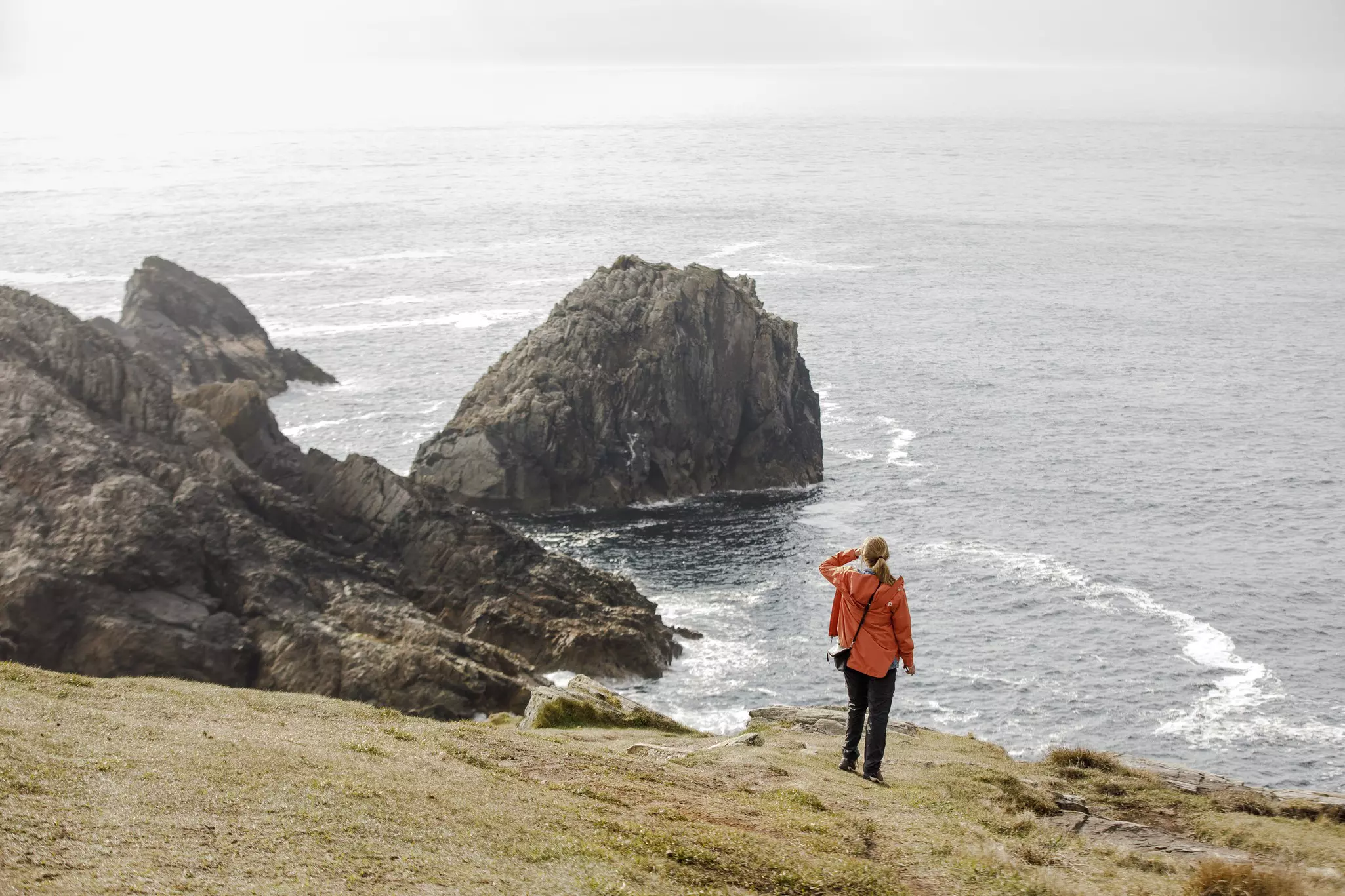 A person in a red jacket on top of a cliff looking at the sea.